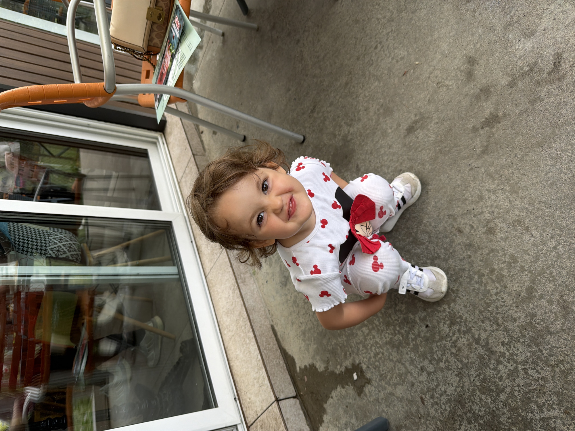 This image features a young child, likely a toddler, crouched on a concrete patio in Plovdiv, Bulgaria. The child, with light brown curly hair and dark eyes, is smiling brightly and looking directly at the camera. They are dressed in a white two-piece outfit patterned with red Mickey Mouse head silhouettes, and white Adidas sneakers with black stripes. A red bow, possibly a Minnie Mouse accessory, is attached to their hip.
The setting is an outdoor seating area, possibly a cafe or restaurant patio. Metal chairs with orange plastic armrests are visible in the background. On one chair, a light-colored handbag featuring a "C" logo (consistent with the Coach brand) rests next to a leaflet that partially displays "COCA COLA" and "Menu." To the left, a large window or glass door with a white frame reflects interior elements, including furniture and cushions. The ground is textured concrete with some damp patches. The lighting suggests it is daytime, with soft, possibly overcast conditions. The child's joyful expression is the prominent emotion conveyed in this candid, high-angle shot.