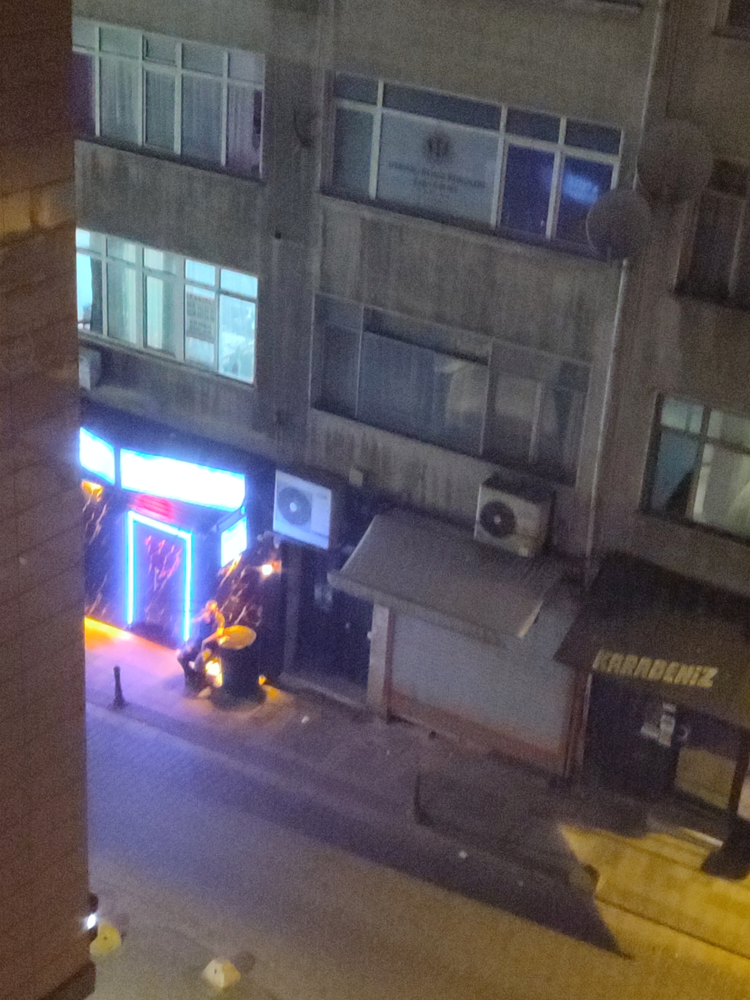 The image depicts a street scene at night in Kadıköy, Turkey. A building facade is prominently featured, with multiple windows visible. The lower section of the building has a business establishment with bright neon blue and red lighting, suggesting a bar or club. One person, appearing to be seated, is visible within the illuminated area, partially obscured by the lighting and possibly by an object such as a barrel. Further to the right, another establishment has a sign with white lettering that reads "KARADENİZ". In the foreground, the street is visible, illuminated by the lights from the businesses. The overall impression is one of a nighttime urban environment with commercial activity.