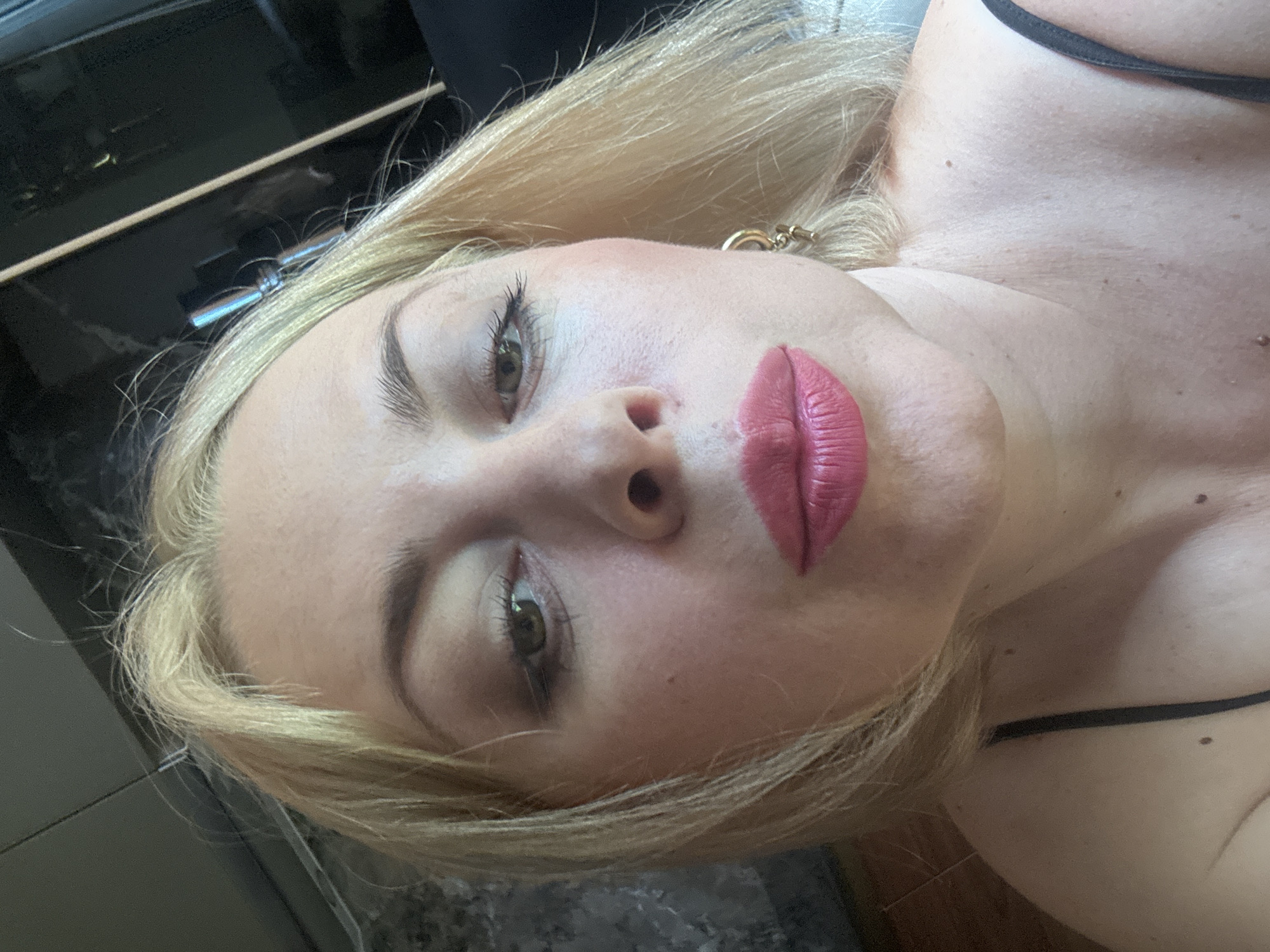 A close-up, high-angle selfie of a blonde woman with fair skin and light brown eyes. She is wearing bright pink lipstick and has on a black spaghetti strap top. She is wearing a gold earring with a small charm. The background is blurred and appears to be an indoor setting. There are no discernible landmarks or details that would indicate the city of Sochi, Russia.