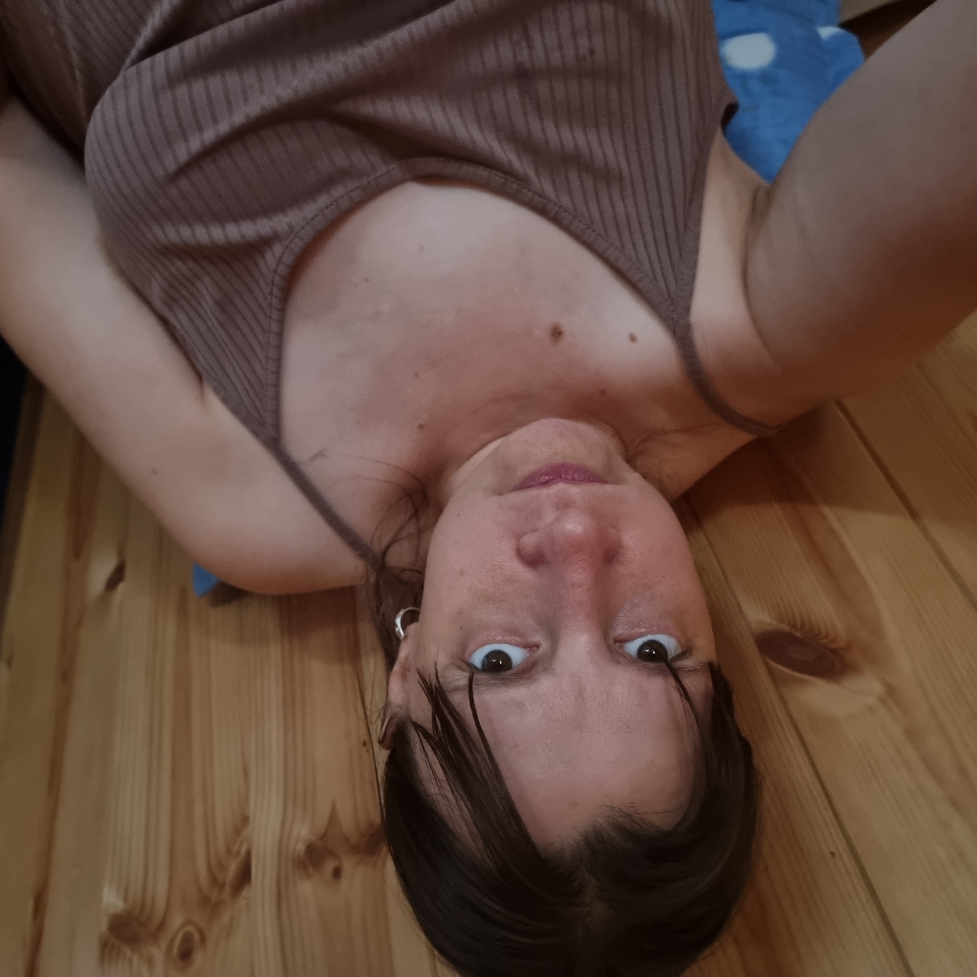 A woman is taking a selfie. She is looking directly at the camera with a slightly displeased expression. She has dark hair that is slightly messy. She is wearing a brown ribbed tank top. The background is a wooden wall. There is a blue object with white polka dots visible in the lower left corner of the image. The image appears to have been taken indoors. The location of Dunnae-myeon, South Korea is not discernible from the image.
