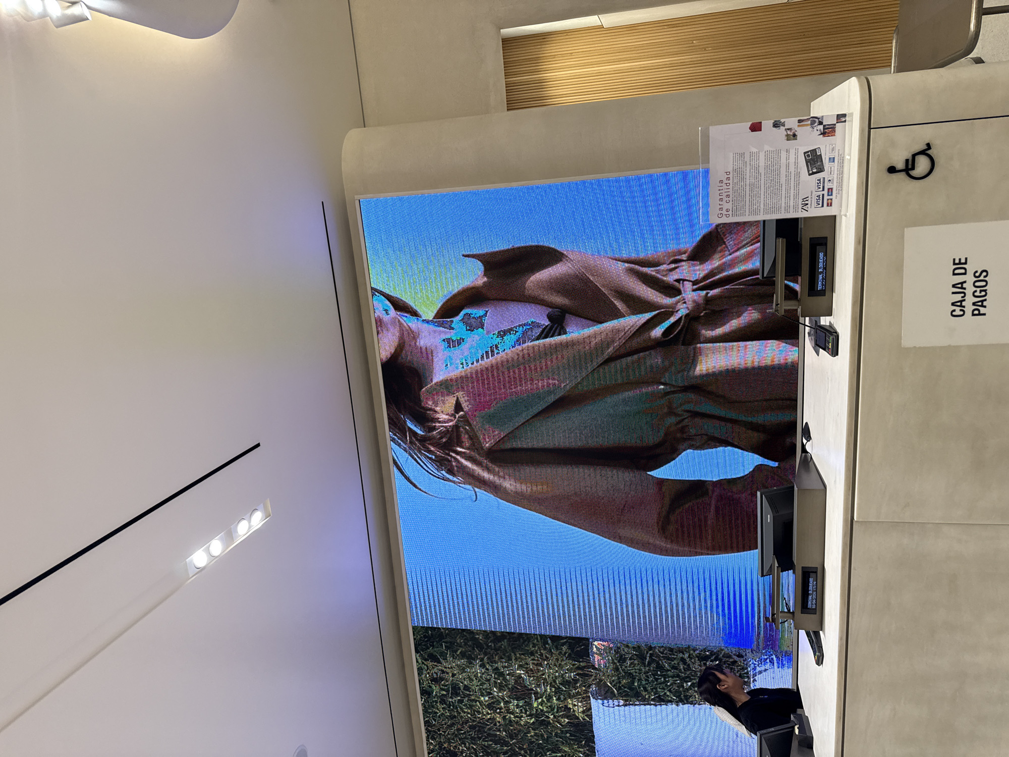 The image depicts a retail environment, possibly a clothing store. A large LED screen displays a fashion advertisement. A checkout counter is visible to the right, featuring a payment terminal, a sign indicating "CAJA DE PAGOS" (Cash Payments), and a sign displaying guarantees of quality and accepted payment methods, including VISA. A person stands near the lower portion of the screen, partially obscured. The setting appears to be indoors with modern lighting fixtures and neutral-toned walls. There is no explicit information in the image to indicate it is located in Puebla, Mexico.