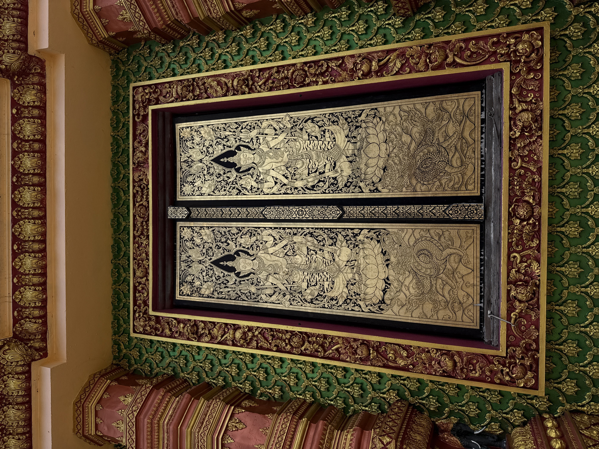 This image captures a richly decorated entrance or window of a building, likely a temple, in the city of Na Nuea, Thailand. The central focus is a pair of ornate golden doors or panels, intricately painted with detailed figures and motifs in black and gold. On each panel, a multi-armed deity stands on a lotus flower, holding various objects, with swirling decorative elements and mythical creatures like dragons below.
The doors are set within a thick, elaborately carved frame. The frame is decorated with a combination of deep red and gold, featuring floral and scrollwork patterns. Surrounding the frame is a textured wall adorned with a repeating pattern of golden motifs on a vibrant green background. The architectural elements extending beyond the frame, particularly on the left and right, are also heavily ornamented with red and gold carvings, including pillar-like structures with tiered designs. The ceiling above is painted in a warm yellow or orange hue with decorative golden trim. The overall impression is one of opulence and intricate craftsmanship characteristic of Thai religious or royal architecture.