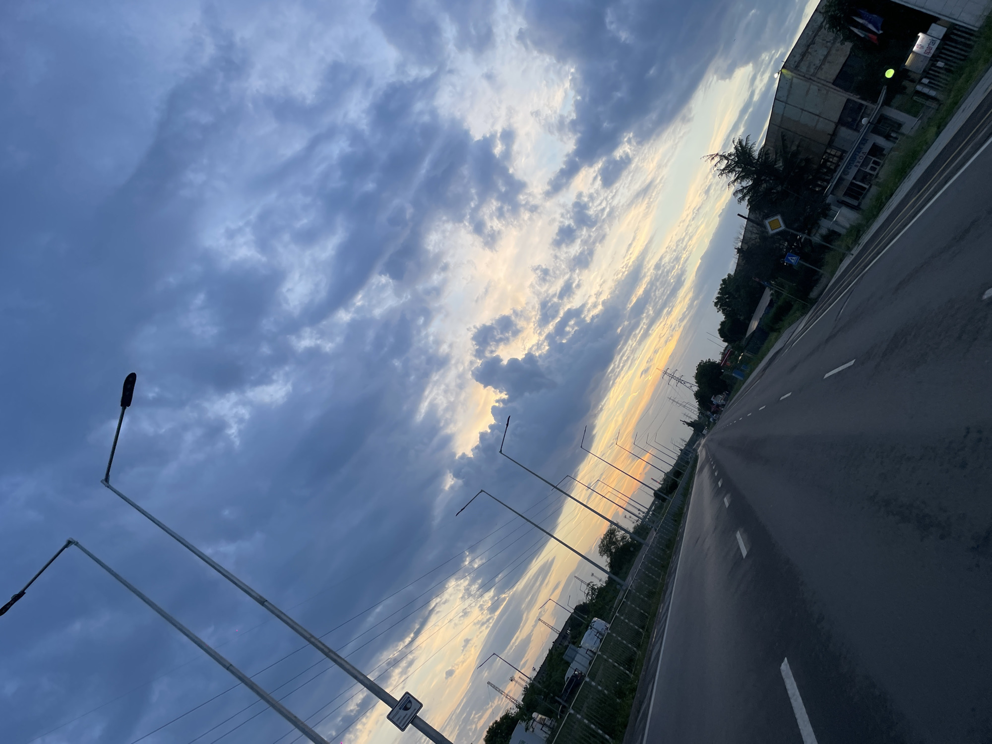 The image depicts a road receding into the distance under a dramatic sky of dark, layered clouds. The sky transitions from blue-gray to a golden sunset hue on the horizon. The road is black asphalt, marked with white lane lines. Streetlights line the left side of the road, receding into the distance. A metal barrier runs along the side of the road. The angle is tilted, giving the impression of a skewed perspective. Vegetation is visible on the sides of the road. A building is visible in the top-right corner, identifiable as the "Sparek" facility. Based on the building and the road infrastructure, it is likely in Burgas, Bulgaria. No people are visible in the shot.