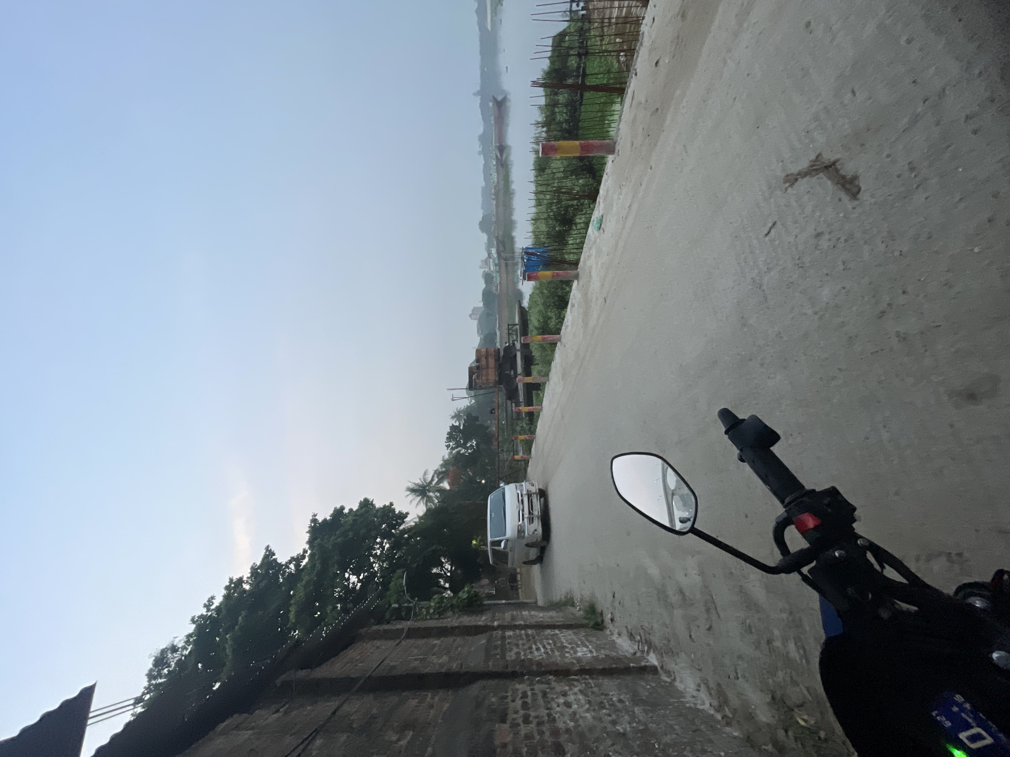 The image shows a view from a motorcycle, likely taken in Dhaka, Bangladesh, on an overcast day. A narrow concrete road stretches into the distance, with a river visible in the background. The riverbank is lined with vegetation and some construction materials. A white van is traveling along the road. On the left, there is a low brick wall and trees. The motorcycle's handlebars and mirror are in the foreground.