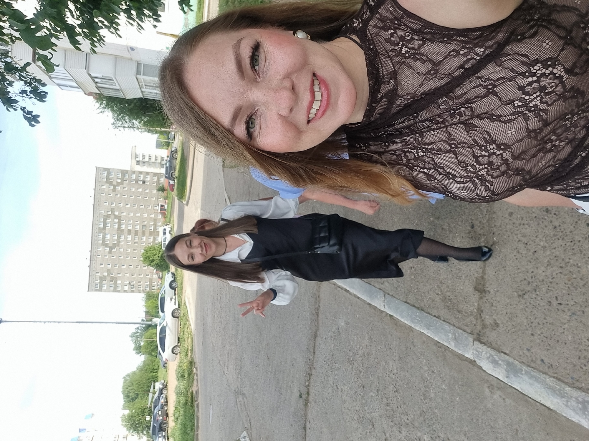 Two women are visible in this outdoor photograph taken in what appears to be a residential area. The woman in the foreground, slightly out of focus, is wearing a lace top. She has light skin, long brown hair, and a smiling expression.
In the mid-ground, the second woman is wearing a black dress over a white blouse with full sleeves, black stockings, and black shoes. She is also light-skinned with long brown hair, and she is holding up two fingers in a peace sign. She appears to be smiling.
The environment consists of a gray asphalt road with a concrete curb. In the background, apartment buildings are visible, as well as parked cars and trees. Based on the architectural style and the presence of Cyrillic characters on some signs in the distance, the location appears to be in a residential area of a city in Russia. Based on the title, the specific location is the city of Chaikovsky, Russia. The sky appears to be partly cloudy.