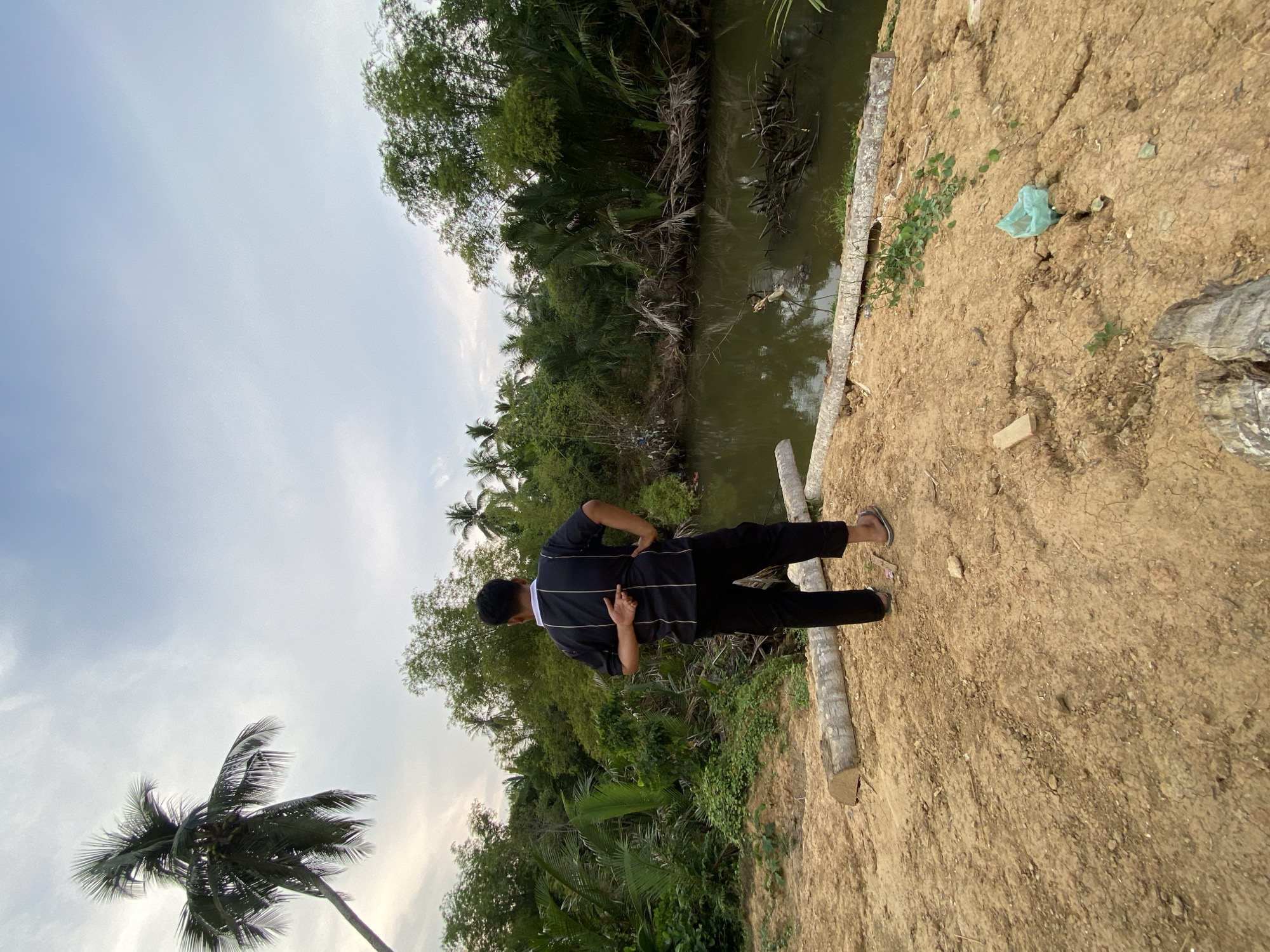 A medium shot captures a man standing on a dirt path next to a body of water, likely a canal or river, in Tanjongkapai, Indonesia. The man has his back to the camera and wears a dark, long-sleeved shirt with light-colored horizontal stripes, black pants, and sandals. He holds his left hand to his back. The vegetation surrounding the water consists of tropical trees, including palms. The sky appears overcast. The ground is dry and cracked. Along the edge of the water, logs are present. A plastic bag sits on the dirt path.