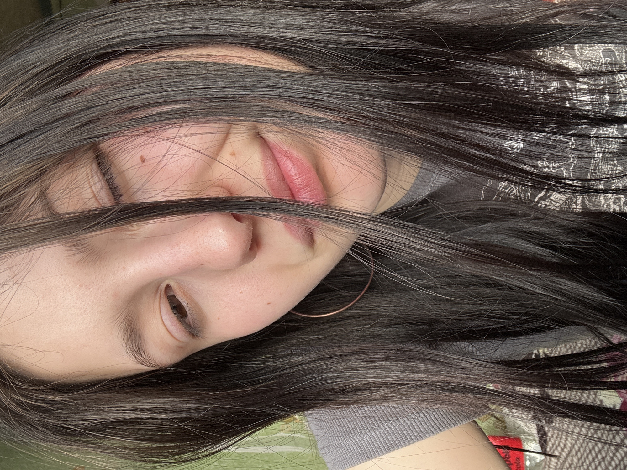 A close-up, slightly rotated, portrait photograph shows a person with dark hair partially obscuring their face. The person's left eye is partially open, while the right eye is closed. The person's lips are visible and slightly parted. A gold-colored hoop earring is visible on the right side of the frame. The person is wearing a gray garment. The background is out of focus, but there is a glimpse of green. The location in the city of Фоки, Russia cannot be determined from the image.