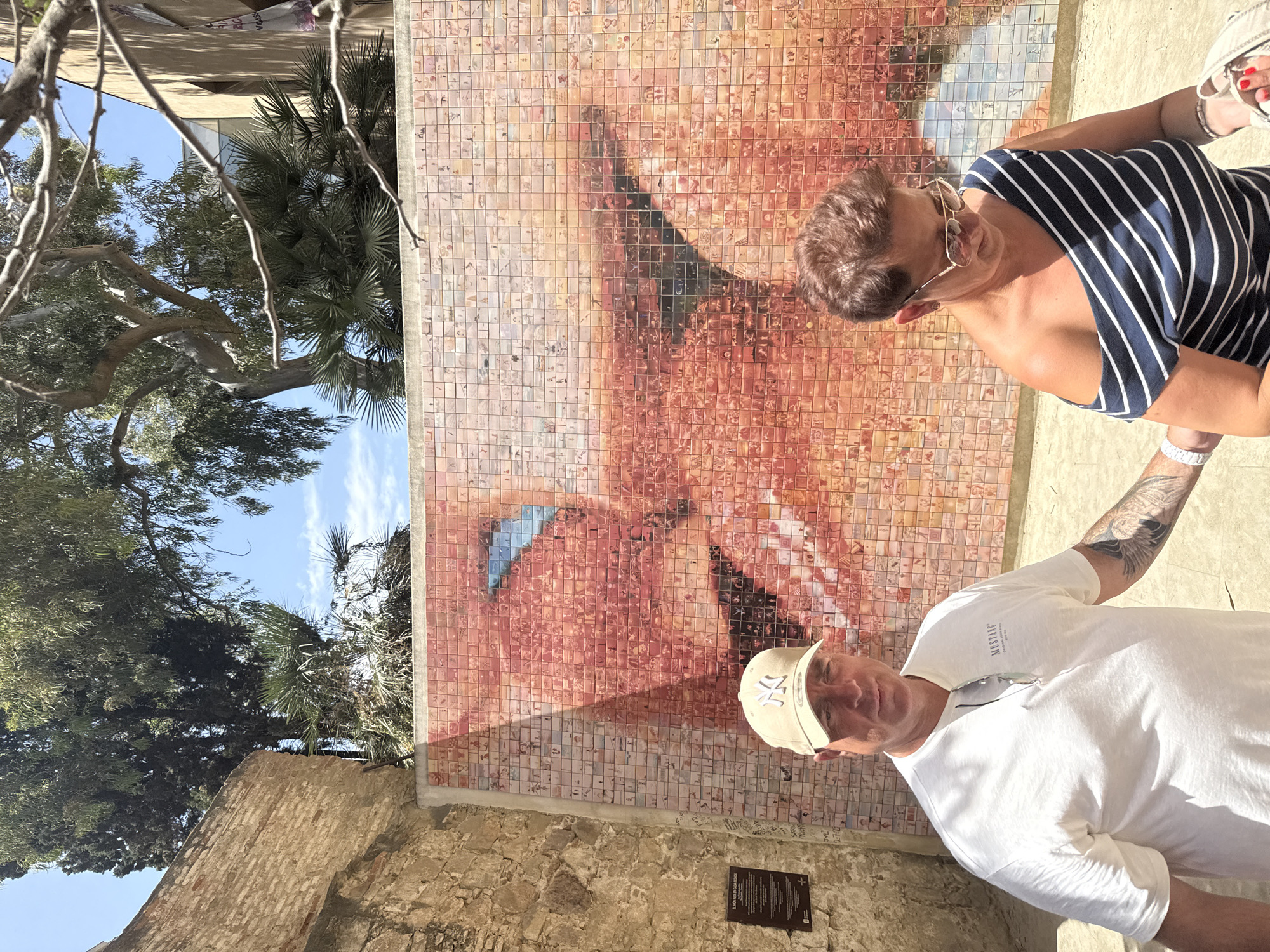 Two people, a man and a woman, are standing in front of a large mosaic mural depicting a woman's face. The mosaic is made of many small square tiles. The woman is wearing a navy and white striped sleeveless top and sunglasses. The man is wearing a white t-shirt and a light-colored baseball cap. He has tattoos on his arm.
The scene appears to be outdoors, with trees and a blue sky visible in the background. The architecture suggests a sunny day, possibly in a historical area. There is some text visible on a plaque on the wall below the mural, but it is not legible in the image. The people's poses suggest they are posing for a photograph. The overall mood appears casual and relaxed. The mosaic itself is a prominent feature, covering a significant portion of the wall.