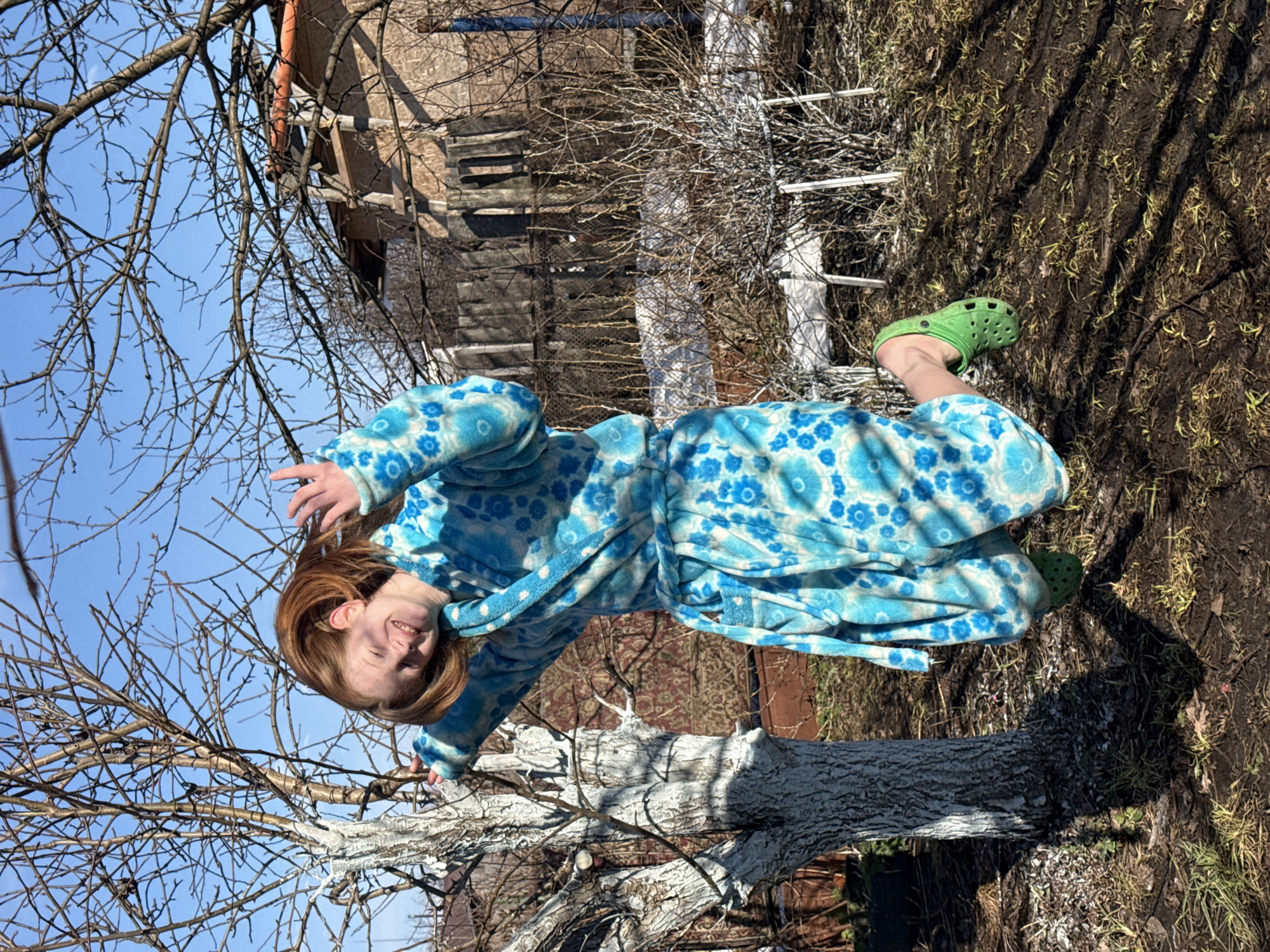 A fair-skinned young woman with reddish-brown hair, wearing a blue and white floral robe and green Crocs shoes, stands outdoors near a tree trunk. The background includes a wooden building with what appears to be a metal roof and bare tree branches against a blue sky. The ground is covered with grass and dirt. The image is rotated approximately 90 degrees clockwise. There is no verifiable information about the location being Chaikovsky, Russia, based solely on visual data.