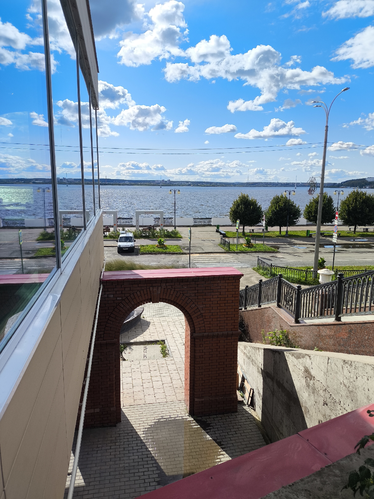 The image depicts a bright, sunny day with a vast blue sky filled with fluffy white clouds. In the foreground, a modern building with large glass windows reflects the sky and the scene beyond. To the right, a brick archway leads to a downward-sloping tiled path and stairs.
Beyond the foreground structures, a waterfront promenade is visible. A wide river stretches across the middle ground, with a distant city skyline on the other side. The promenade is lined with trees, lampposts, benches, and decorative black metal railings. A white van is parked on the promenade, and a few people are seated on benches. The overall environment suggests a public space, possibly a park or embankment, in an urban setting.
The view is consistent with the embankment of the Izhevsk Pond in Izhevsk, Russia, specifically near the monument to the founders of Izhevsk. The architecture of the buildings and the style of the promenade are characteristic of this location. The presence of the pond and the distant cityscape further support this identification.