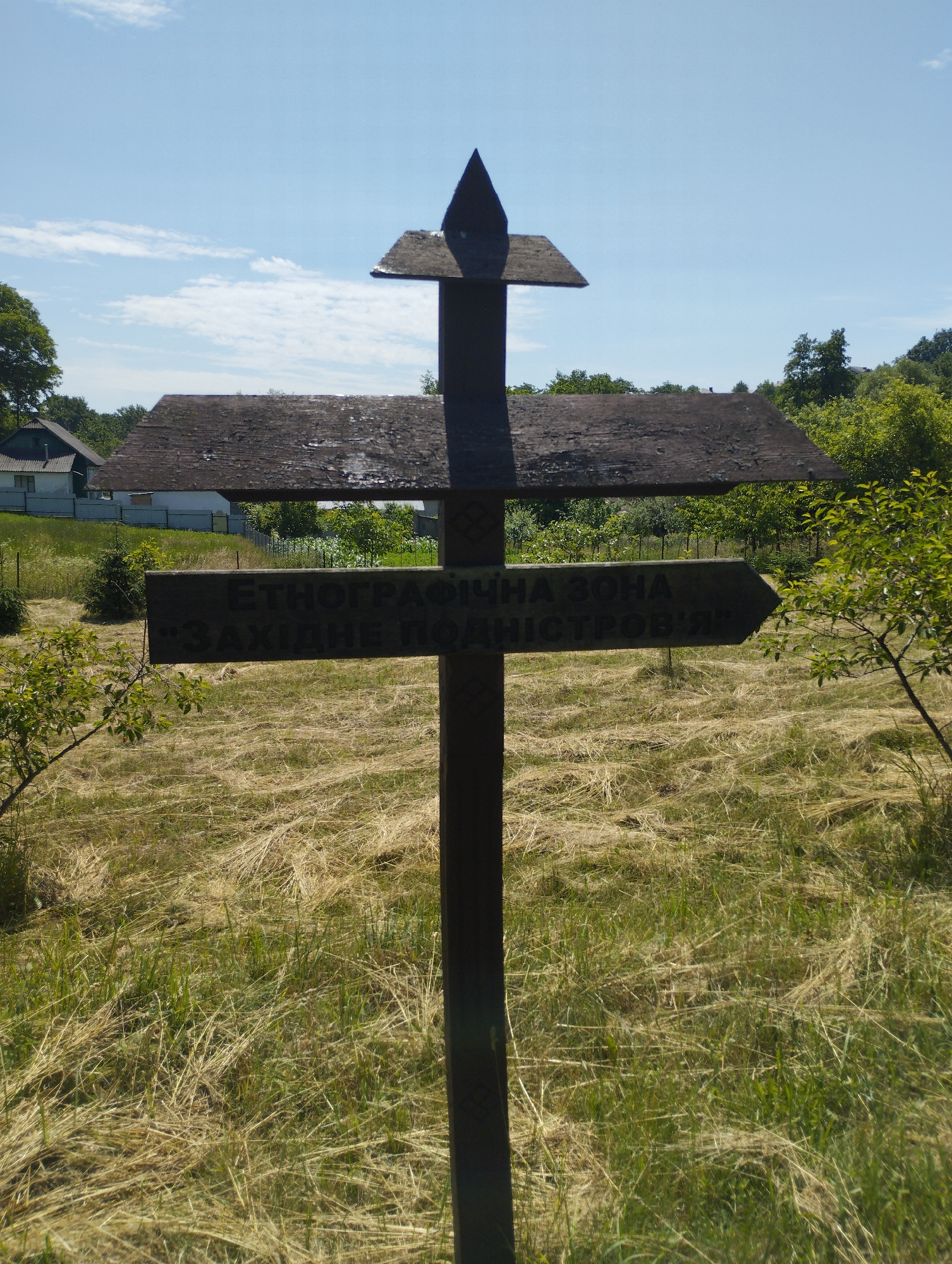 A vertical, wooden cross-shaped sign stands in a grassy field under a bright blue sky with scattered clouds. The sign has a pointed top and two horizontal beams. The upper beam is shorter and points upwards, while the lower, longer beam is shaped like an arrow and points to the right. The lower beam has text inscribed on it in Cyrillic letters. Behind the sign, two buildings with dark, pitched roofs are visible, along with a fence and green trees. The field is covered in dry, yellow grass and some green vegetation. The sign appears to be weathered.