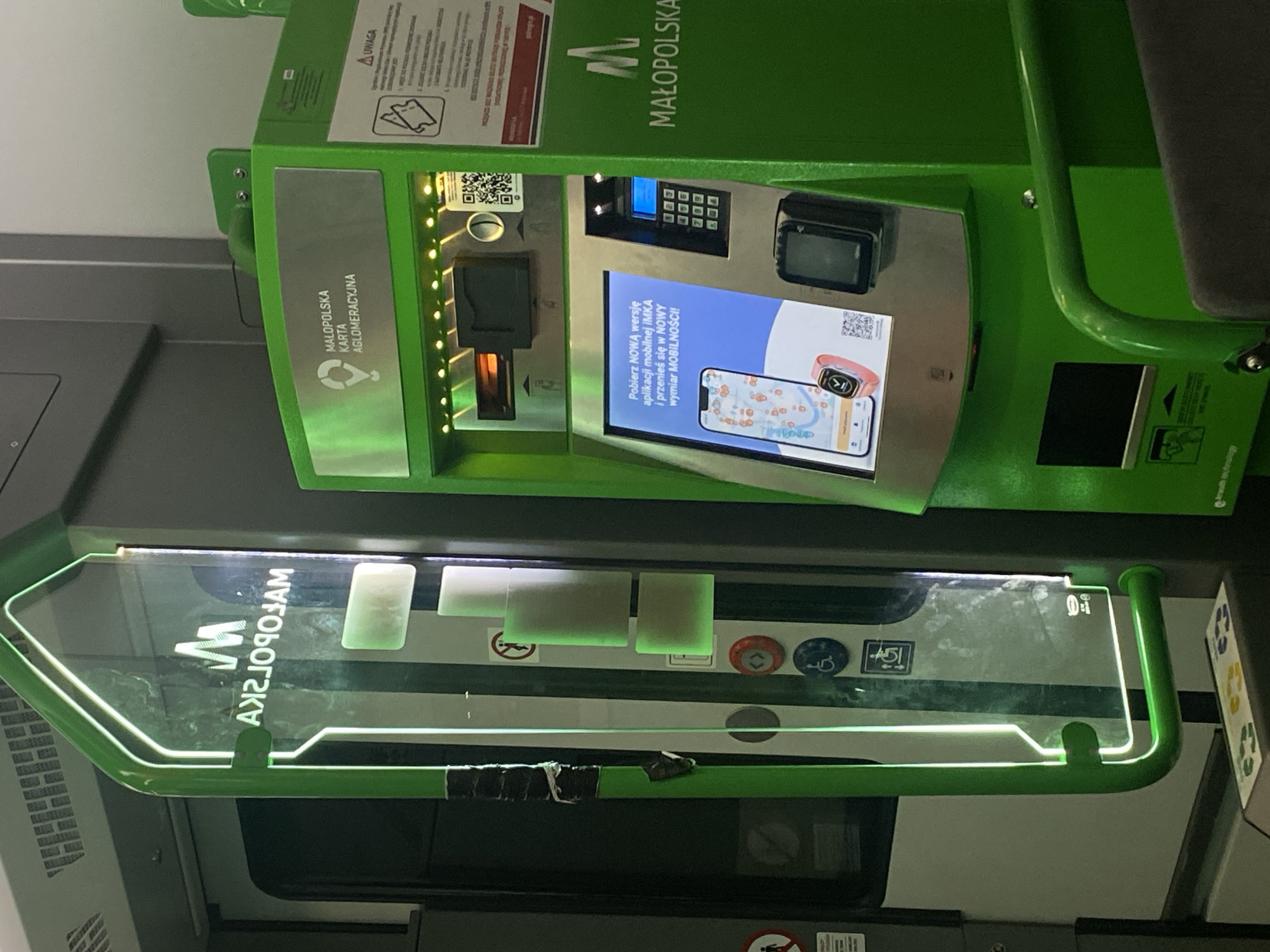 This is a photograph of a ticket vending machine and a vertical advertising display, both in bright green, placed side-by-side. The vending machine features a touch screen displaying an advertisement for a mobile application, a keypad for entering information, a card reader, and a slot for tickets. Above the screen, there are slots for inserting cash and a QR code. A logo with the letters "M" and "MAŁOPOLSKA" is visible on the machine.
The vertical display to the left is illuminated with white light and has the same "M" logo and "MAŁOPOLSKA" text, but reflected and reversed due to the display's surface. Below this text on the display are several rectangular, softly lit green panels. Further down on the side of the vertical display, there are icons for accessibility and a circular button with an arrow symbol.
The background appears to be the interior of a public transportation vehicle, possibly a train or bus, with grey walls and seating visible. On the lower part of the vending machine, there is a green handrail. A small sign with recycling symbols is visible at the bottom of the image.
While the image itself does not provide explicit confirmation of the location being Rzuchowa, Poland, the prominently displayed text "MAŁOPOLSKA" refers to the Lesser Poland Voivodeship, a region in southern Poland where Rzuchowa is situated. Therefore, it is highly probable that this scene is located within Rzuchowa, Poland, likely at a public transportation hub or on a vehicle serving the region. No individuals are present in the frame. The environment appears to be clean and functional.