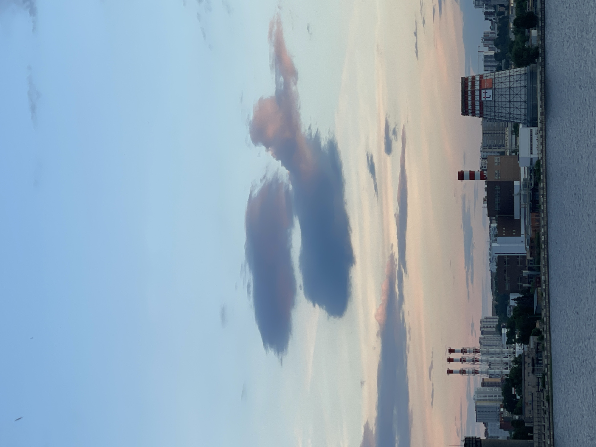 The image depicts an urban or industrial landscape at what appears to be either sunrise or sunset, given the soft, gradient sky. The scene is dominated by a vast sky that transitions from a light blue at the top to a pale orange and pink hue near the horizon. Scattered across the sky are various cloud formations, including several distinct, dark, and somewhat clumpy clouds, and lighter, wispy streaks. Notably, one of the darker cloud formations bears a striking resemblance to a human figure with arms raised overhead, creating a vivid example of pareidolia.
Below the expansive sky, along the horizon, lies a built-up area adjacent to a calm body of water, likely a river or canal, which occupies the rightmost portion of the frame. The cityscape features various buildings and industrial structures. A prominent feature is a large, lattice-like industrial tower with red and white striped accents near its top, located towards the right. On its side, a large rectangular sign is visible, displaying the text "МОСКОВСКИЙ ТЕПЛОВОЙ" (Moskovskiy Teplovoy, meaning "Moscow Heat Power") and the number "+7". Further to the left, several slender, red and white striped chimneys or smokestacks rise from other industrial facilities. Various other buildings, ranging from multi-story structures to what appear to be residential blocks, complete the urban skyline. The overall atmosphere is calm and serene, with the soft light of the early morning or late evening.