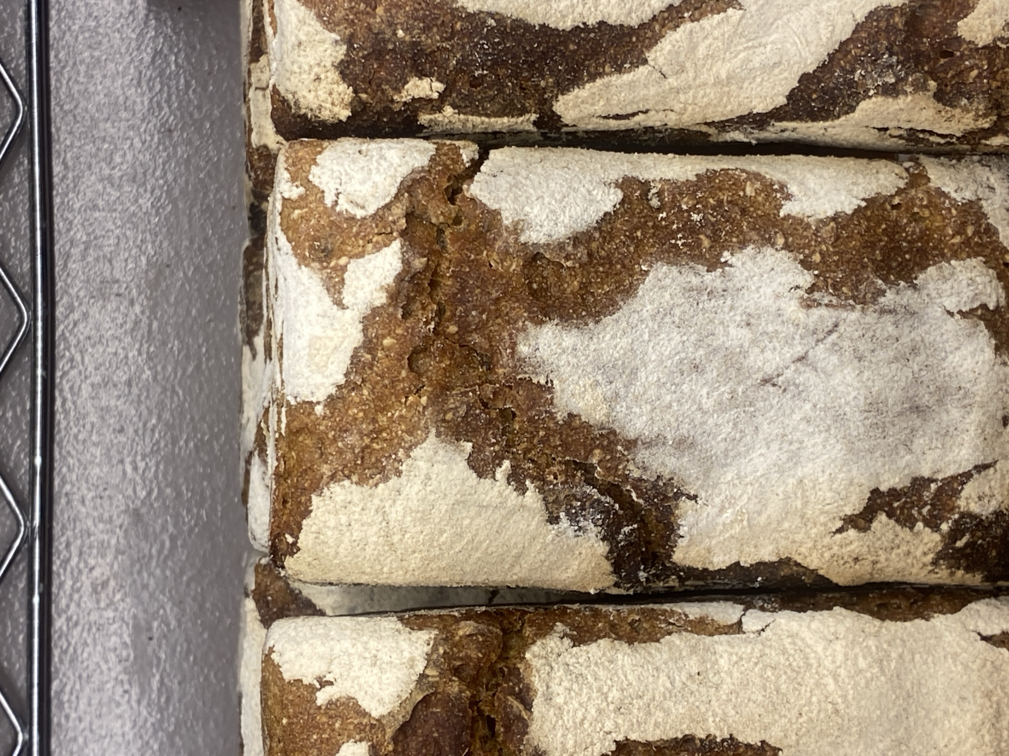 The image shows a close-up, overhead view of three loaves of dark, rustic bread stacked on top of each other. The bread appears to be rye bread, characterized by its dark color and the visible grains and seeds within the crumb. The crust is a deep brown, with irregular patches of white flour dusted across the surface. The texture of the bread is rough and hearty, suggesting a crusty exterior and a dense interior. The loaves are tightly packed, with a slight gap visible between the top two loaves and the bottom two. To the left of the bread, a portion of a wire rack is visible, suggesting the bread is being stored or displayed on a cooling rack or shelf. The background is a textured, light gray surface, possibly a wall or countertop. There is no visible text in the image, and no specific time of day or weather conditions can be inferred. The setting is likely a bakery or a place where bread is sold. The focus is entirely on the bread itself, highlighting its artisanal appearance.