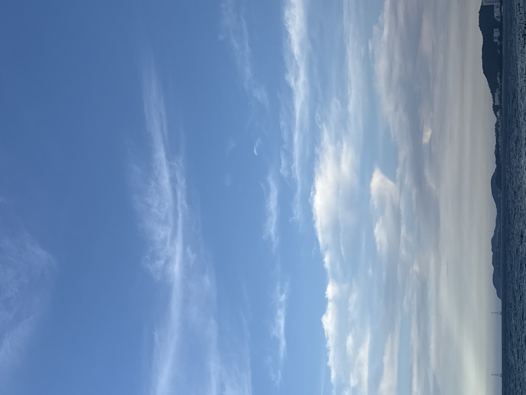 This is a landscape photograph taken from a body of water, likely the sea, looking towards a coastline. The dominant subject is the sky, which occupies the majority of the frame and displays a mix of clear blue and wispy white clouds. The clouds are thin and streaky in some areas, with thicker, more substantial formations towards the right side of the image.
On the right side of the image, a silhouetted coastline is visible, characterized by a dark, mountainous terrain. The outline of buildings is discernible on the slopes of the hills, suggesting an urban or semi-urban settlement. The sea in the foreground is a deep, dark blue, with subtle ripples indicating a calm to moderate water condition. Two slender, tower-like structures, possibly lights or communication towers, rise from the water in the distance along the coastline.
The image appears to be taken during the daytime, with the bright sky and diffused lighting suggesting either morning or late afternoon. The weather seems clear with scattered clouds. The presence of the sea and the coastline with buildings points to a coastal location, specifically identified as Nha Trang Ward, Vietnam. There is no visible text in the image. The overall mood of the photograph is serene and expansive, focusing on the natural beauty of the sky and the seascape.