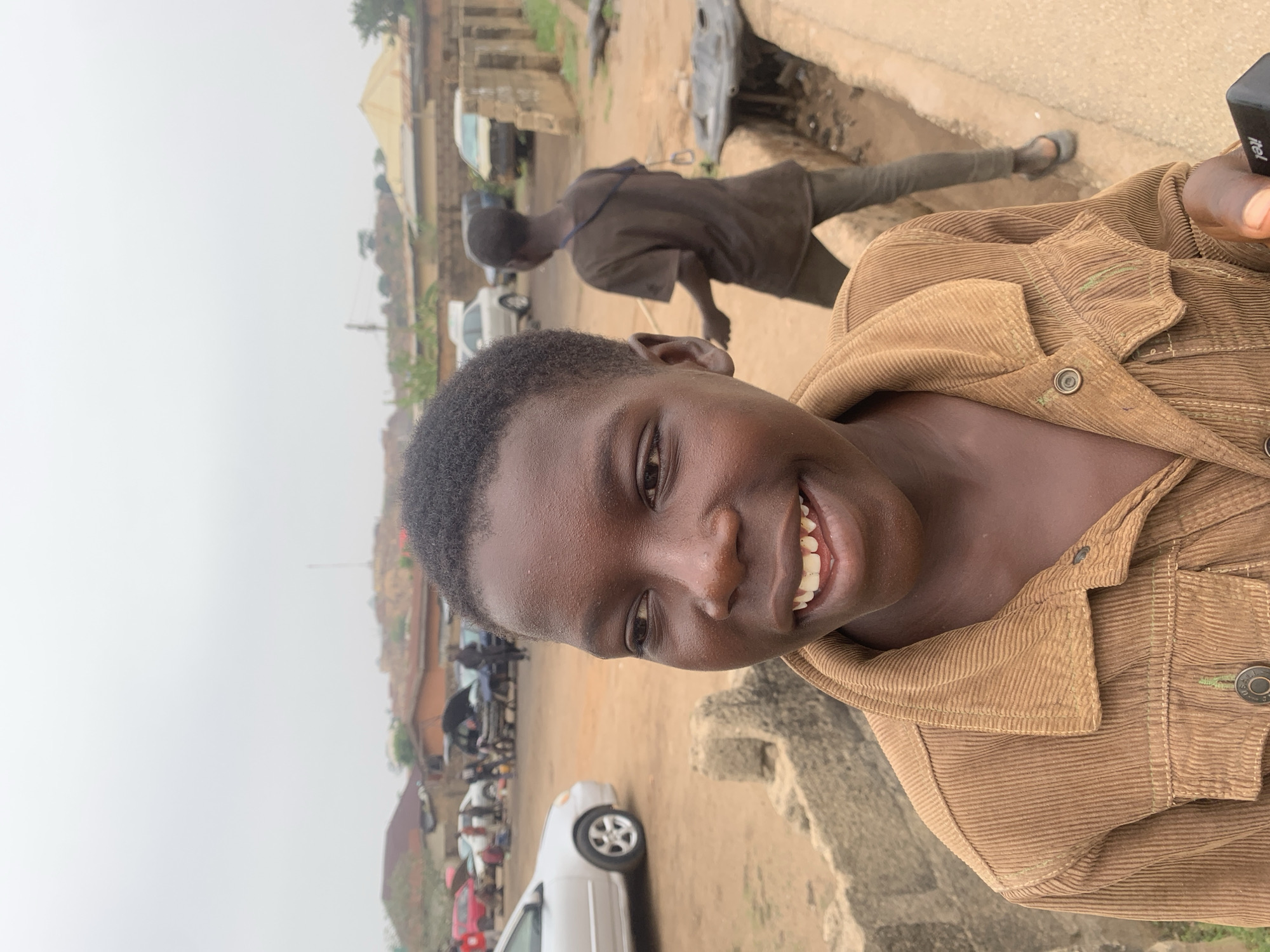 A young person with dark skin and short, dark hair is smiling at the camera while wearing a brown corduroy jacket. The background features a somewhat cluttered urban scene, including buildings, parked cars (white and red), and other people. Another young person is crouched near what appears to be an open drain or gutter. The location is Jos, Nigeria.