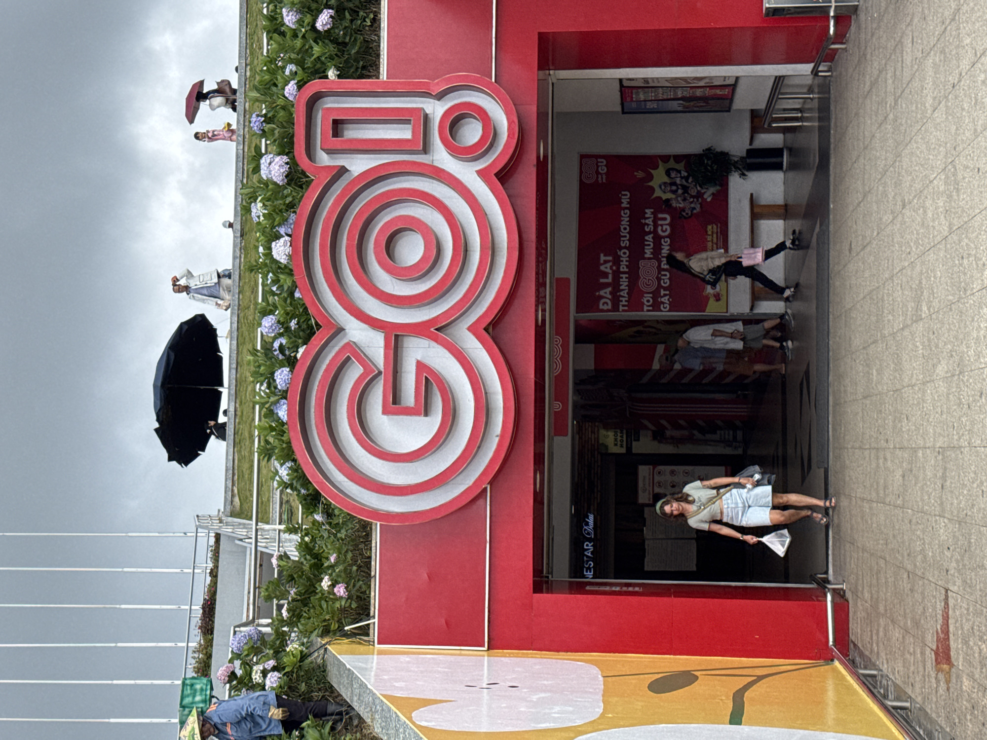 This image captures an outdoor scene at a commercial entrance in Da Lat, Vietnam, under an overcast sky. A large, prominent red and white sign displaying the word "GO!" stands in the foreground, featuring a stylized, almost 3D design. To the left of this sign, a garden bed is filled with blooming hydrangeas in shades of blue, purple, and white. Beyond the garden, several people are visible, some holding umbrellas (one black, one red), suggesting damp or rainy weather. One individual is seen wearing a blue work uniform and a straw hat.
To the right of the large "GO!" sign is the entrance to a building, likely a store, with glass doors. Inside, vibrant red advertising posters are visible, bearing Vietnamese text. Key phrases on these posters include "ĐÀ LẠT" (Da Lat), "THÀNH PHỐ SƯƠNG MÙ" (City of Fog), and promotional messages like "TỚI GO! MUA SẮM GẬT GÙ ĐÚNG GU" (Come to Go! Shopping, perfectly matching your style), with the brand "GU" also displayed. Several people are inside the entrance, including a woman in a light top and shorts who is looking towards the camera, holding a small white net or bag. Another person is seen further inside, walking into the store. A partial view of a yellow and white graphic is visible on the ground in the immediate foreground. The scene depicts typical activity around a retail space during what appears to be an overcast day.