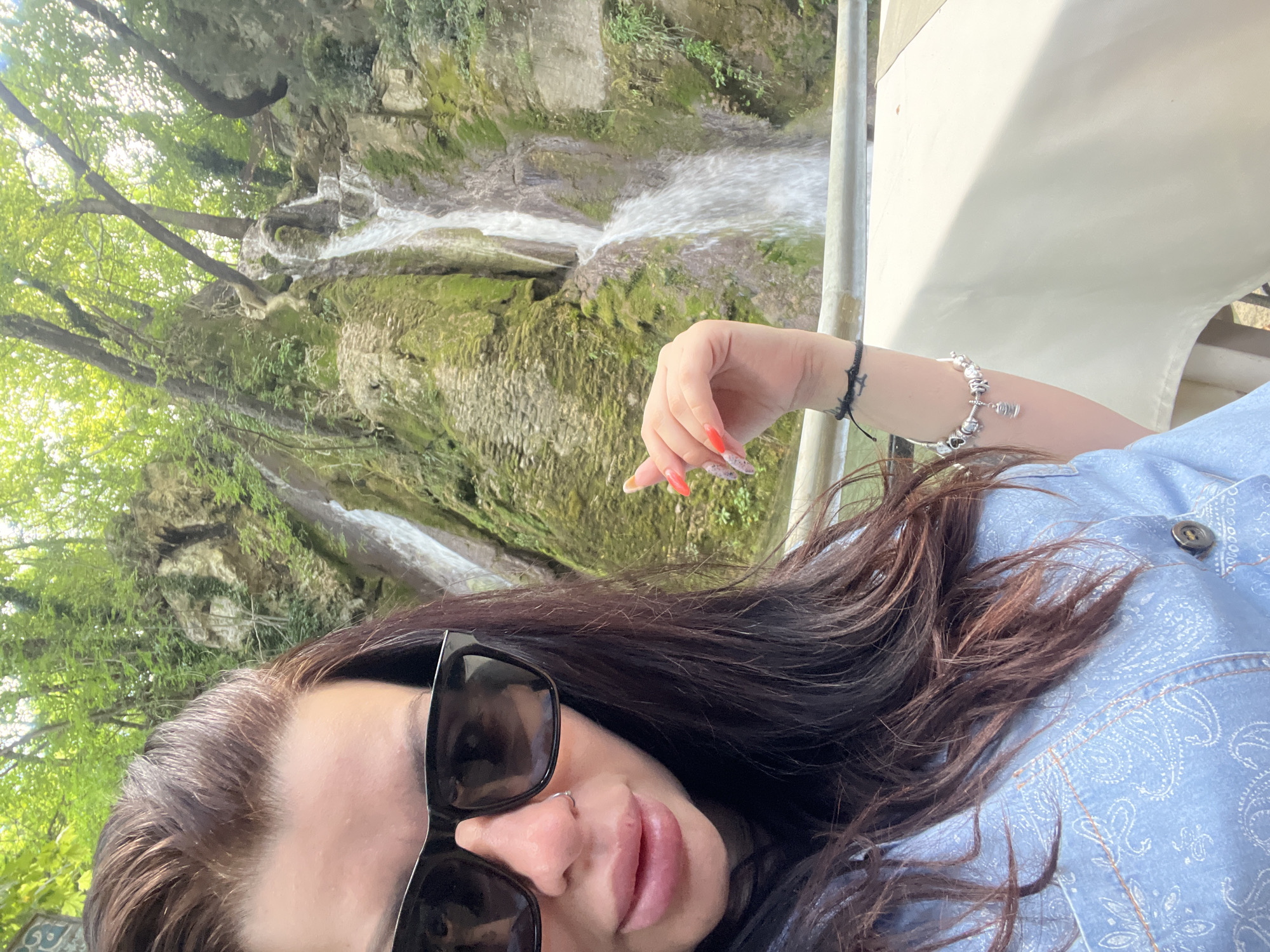 In the city of Bachkovo, Bulgaria, a woman with dark brown hair and sunglasses takes a selfie in front of a waterfall. She is wearing a light blue denim jacket and her right hand, with long, pointed, red and white-patterned nails, is resting on a railing. A bracelet with multiple charms adorns her wrist. The waterfall cascades down a moss-covered rocky cliff face, surrounded by lush green trees and vegetation. The scene suggests an outdoor natural setting, possibly a park or a tourist attraction.