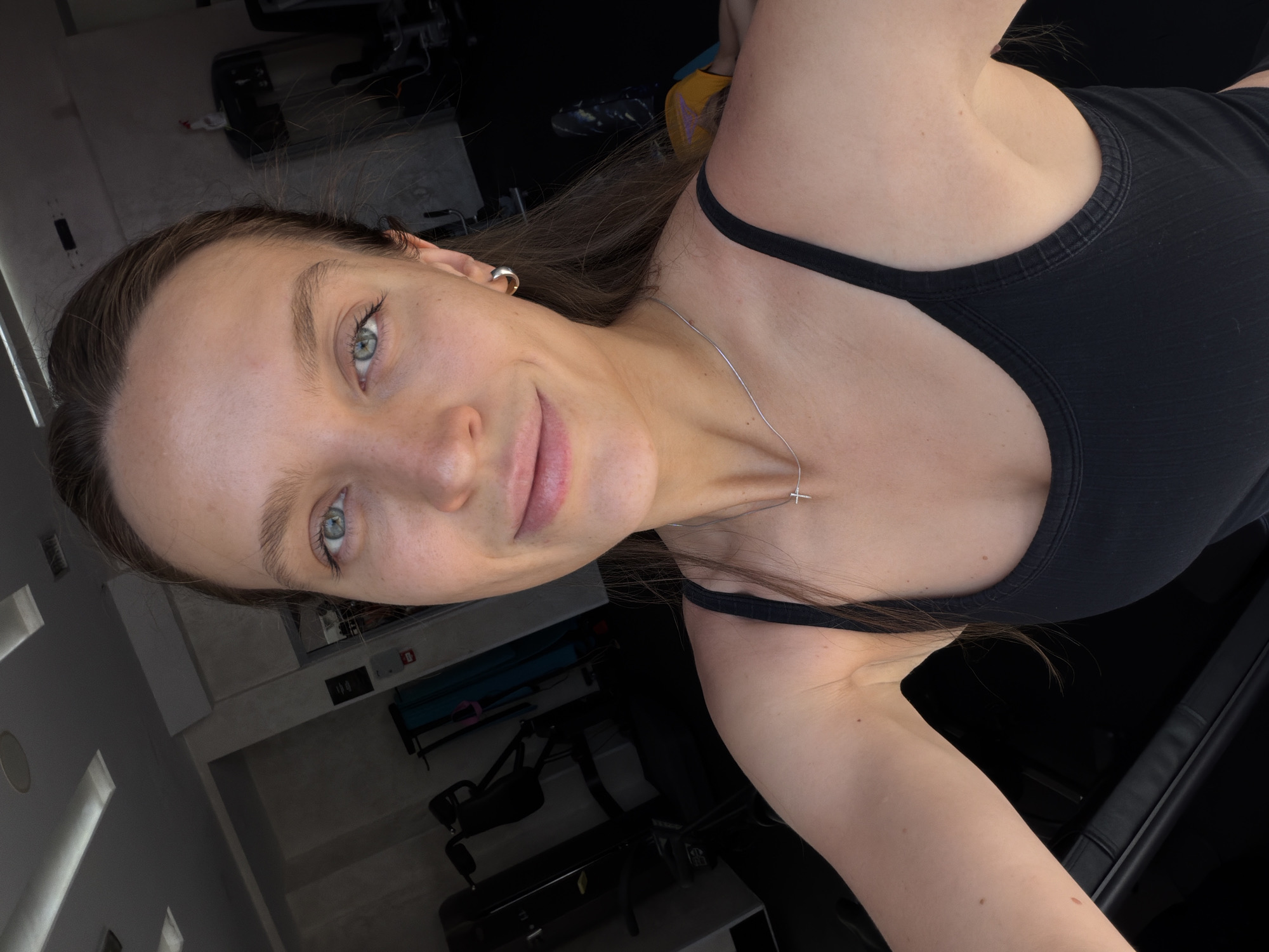An outdoor, medium shot captures a woman with light brown hair and blue eyes. She is smiling and looking directly at the camera. She is wearing a black sports bra and a silver necklace with a cross pendant. The background appears to be a gym with exercise equipment visible. The lighting suggests it is daytime. The location is Budva, Montenegro.