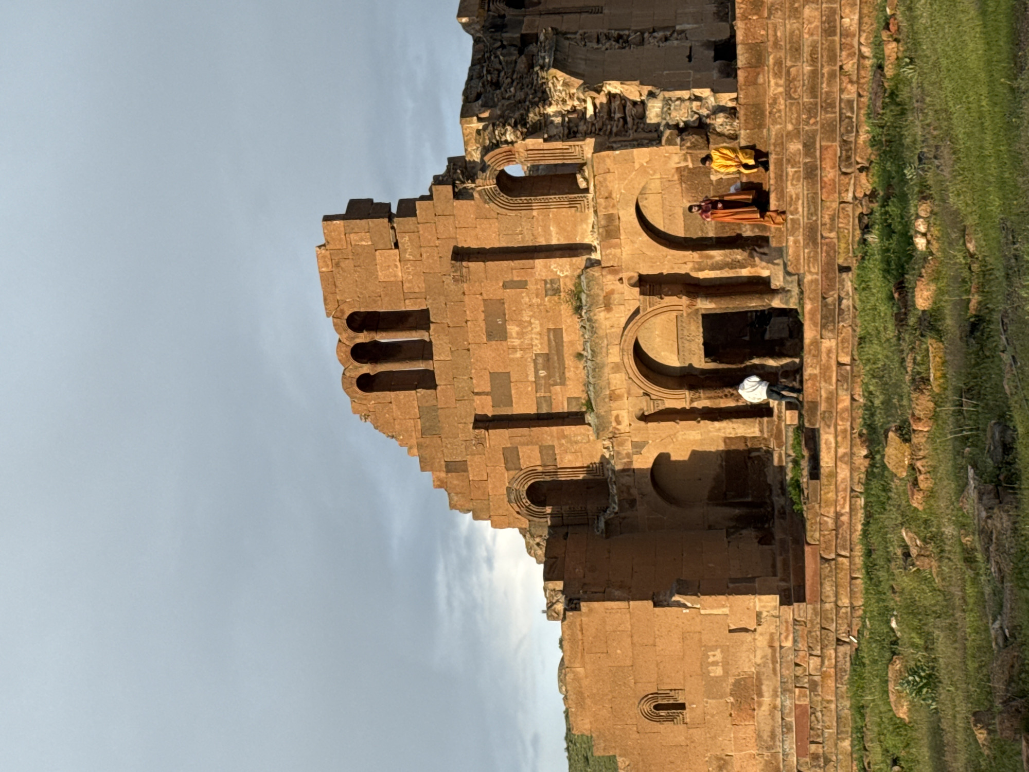 The image depicts a section of the ruins of the Cathedral of Ani in Anipemza, Armenia. The structure is made of tan stone, with a pattern of arches and rectangular recesses in the wall. Two people in robes are standing on the edge of the structure, and a third person in a white shirt is kneeling. The background includes a blue sky with some clouds and green grass in the foreground.