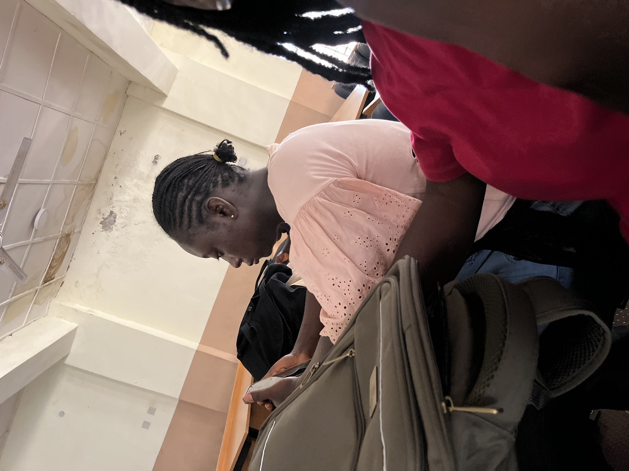 In a classroom setting, possibly in Jos, Nigeria, a young Black woman with braided hair styled in cornrows and pulled back into a bun is seen from the chest up, looking at her phone. She is wearing a light pink blouse with eyelet details. In the foreground, to her left, is a gray backpack with a gold zipper pull. To her right, partially visible, is another person wearing a bright pink garment. The background reveals classroom walls with beige and brown accents and a ceiling with water stains and a fan. Desks are visible behind the subjects.