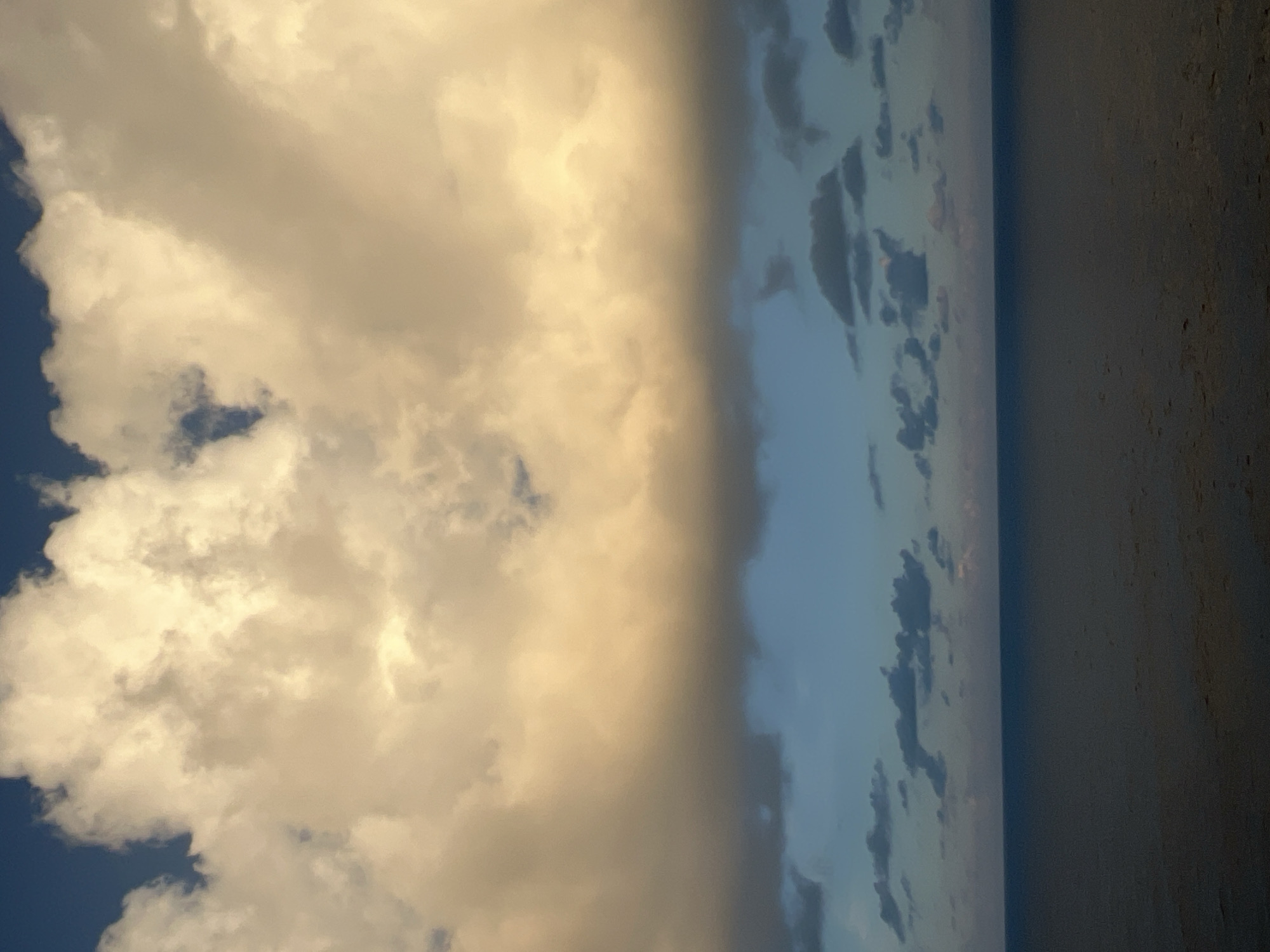 This is a photograph of the sea and sky. The sky is filled with clouds, with a large band of yellowish-orange clouds across the middle and darker blue sky above and below. The sea is dark and appears calm, with subtle ripples on the surface. The image is taken from a high vantage point, looking out over the water.