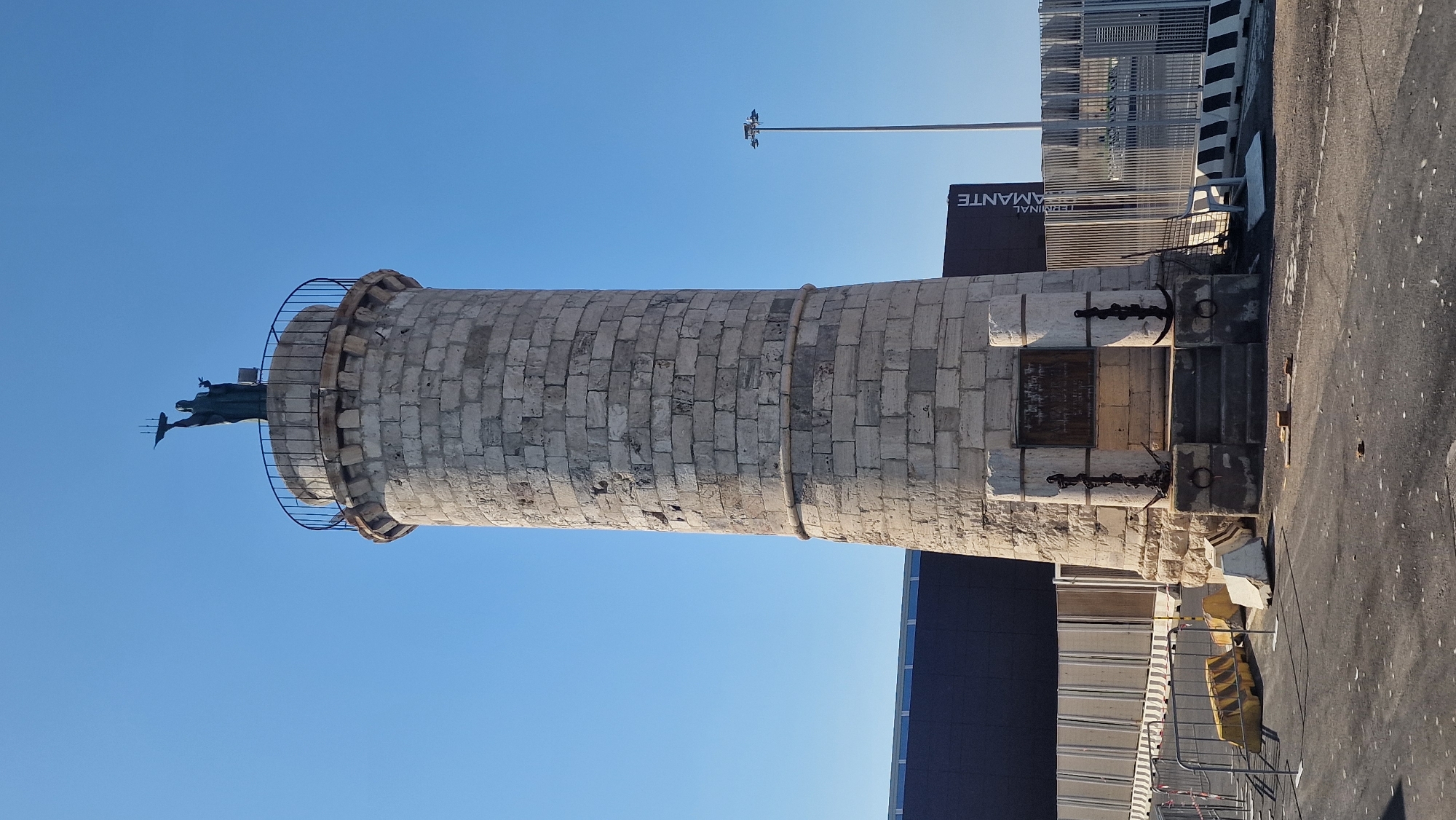 This is a full-shot, eye-level, outdoor, daylight photograph of a stone lighthouse in the city of Civitavecchia, Italy. The lighthouse is cylindrical and made of rough-hewn, light-colored stones. It has a metal railing around the top, where a dark statue of a person holding a ship is perched. The base of the lighthouse is wider and has two stone pillars on either side of a set of stairs. Each pillar has an anchor attached with a chain. There is a plaque on the lighthouse wall above the stairs.
Behind the lighthouse, a dark building with a blue stripe is visible, and further back, a tall light pole stands against a clear blue sky. Metal fencing and concrete barriers are present in the foreground and to the right of the lighthouse. The ground is asphalt, with some yellow and white traffic barriers and loose bricks scattered around. A sign that reads "TERMINAL AMANTE" is visible on the dark building.
