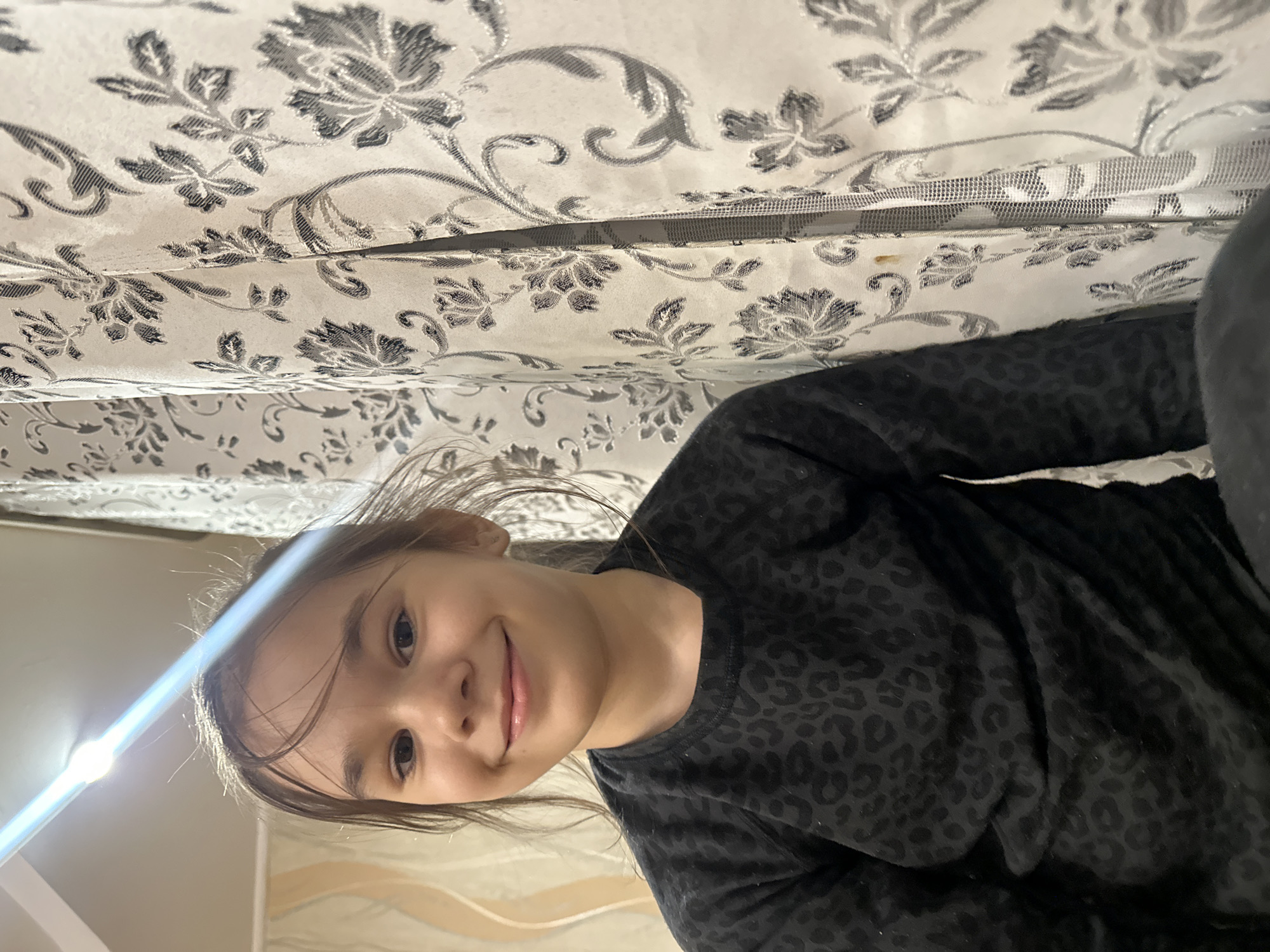 The image is oriented sideways, appearing rotated 90 degrees clockwise. It features a young girl, likely pre-teen, looking upwards with a gentle, cheerful smile directed at the camera. Her light brown hair is loosely pulled back, with a few strands framing her face. She is wearing a dark, possibly black or very dark grey, long-sleeved top featuring a subtle, tone-on-tone leopard print pattern.
Above and slightly behind her, a light-colored fabric with a repeating grey and white floral pattern is prominent, suggesting curtains or bedding. Detailed crops reveal that this fabric might include a mesh layer beneath the patterned material. A strong, bright light source is visible to the girl's left, creating a significant lens flare and illuminating her face. The overall setting appears to be indoors, likely a home environment. No visible text is present in the image. The girl's expression conveys a sense of happiness and ease as she poses for the photograph.