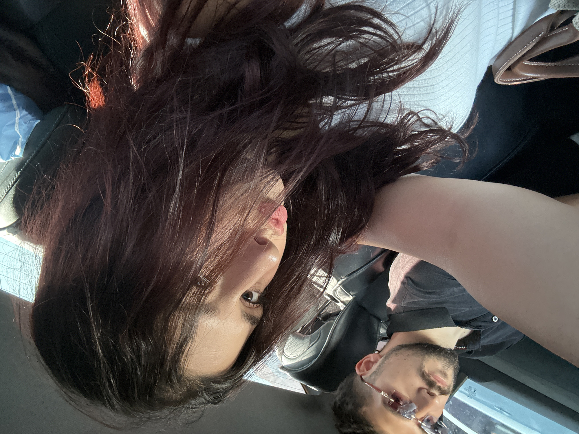 In the city of Baku, Azerbaijan, two individuals are seated in the back of a car. A man, wearing a black polo shirt and rimless sunglasses with a pink tint, is positioned in the driver's seat, his gaze directed slightly away from the camera. Beside him, a woman with long, dark auburn hair is leaning towards the camera, her face partially obscured by her hair. She is wearing a white, textured top. Her left arm is extended, appearing to hold or support the camera, and her lips are slightly parted. The interior of the car is visible, with black leather seats and a light-colored headliner. The sun shines through the rear window, casting a warm glow on the scene. A brown handbag rests on the seat in the foreground, and a blue plastic bag is visible in the background.