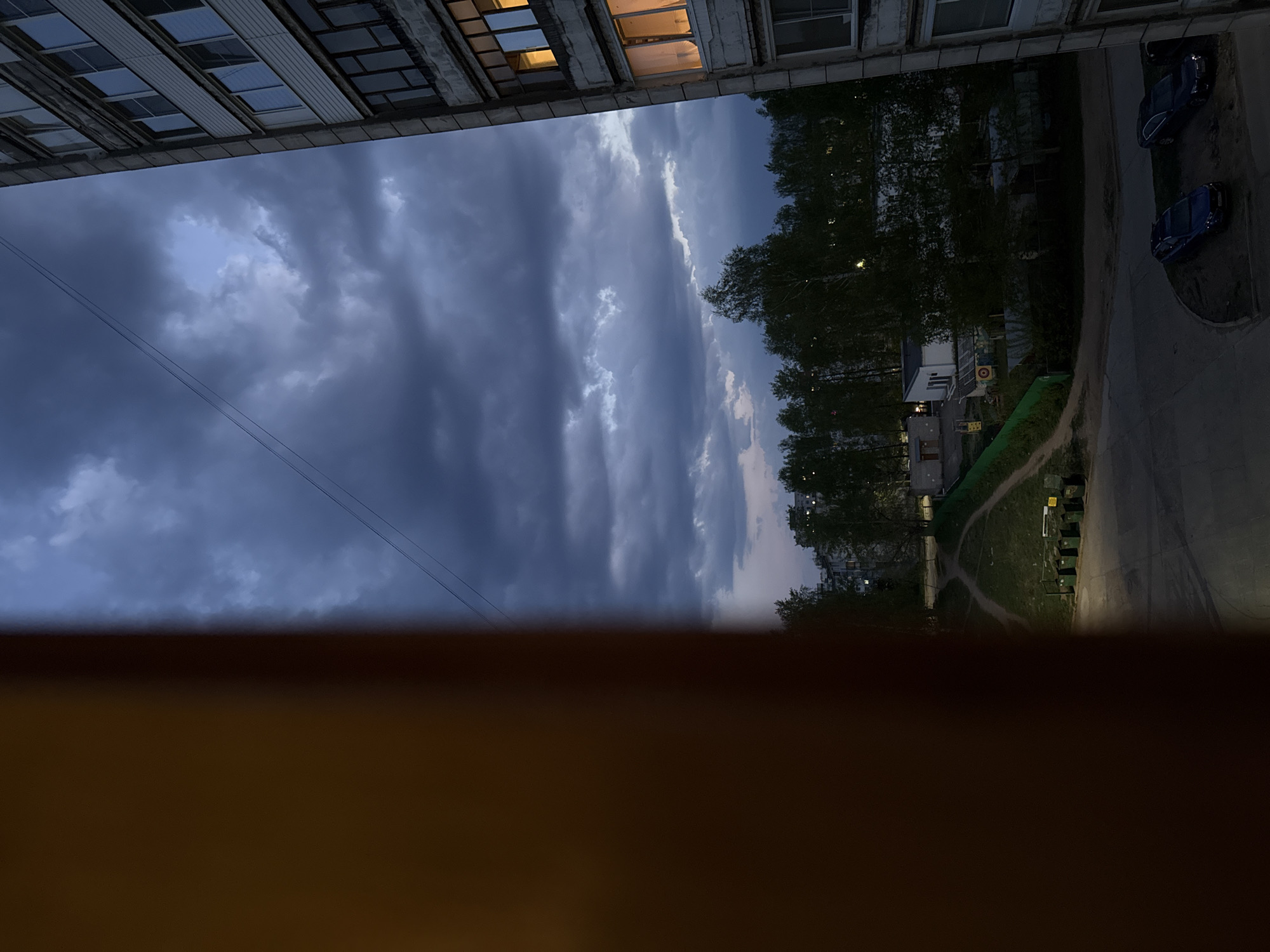 The image depicts an urban scene at dusk, possibly viewed from a balcony. Two apartment buildings are visible, with a cloudy sky overhead. A green area with trees and a sidewalk is situated between the buildings. A road runs adjacent to the green space, where two parked cars are visible. Several refuse bins are on the edge of the sidewalk. The location is likely in an urban environment with residential buildings.