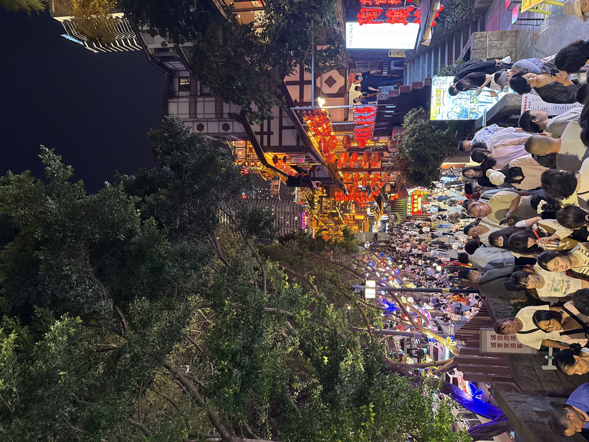 This image captures a vibrant and densely crowded pedestrian street or night market in Yuzhong District, China, at night. The scene is illuminated by numerous artificial light sources, creating a lively and festive atmosphere.
Hundreds of people, dressed in casual clothing, fill the thoroughfare, walking, standing, and socializing. The crowd is particularly dense in the middle and far distance of the street. Traditional Chinese architecture lines both sides of the street, characterized by multi-story buildings with dark tiled roofs, white walls, and visible wooden structures, some adorned with decorative patterns.
Overhead, countless red paper lanterns are strung across the street, casting a warm, inviting glow. Additionally, tree branches lining the street are decorated with colorful lights shaped like butterflies and flowers. Lush green trees with dense foliage are prominent in the foreground and along the street, their leaves illuminated by the ambient lights.
Various signs with Chinese characters are visible throughout the scene. On the right side, a red sign clearly displays "请勿攀爬" (Please do not climb) and "禁止吸烟" (No smoking). Another illuminated sign in the middle ground features cartoon figures and Chinese text. The dark sky confirms the nighttime setting, and the abundance of lights emphasizes the street's activity after dusk. The scene conveys a sense of bustling urban life and cultural engagement.