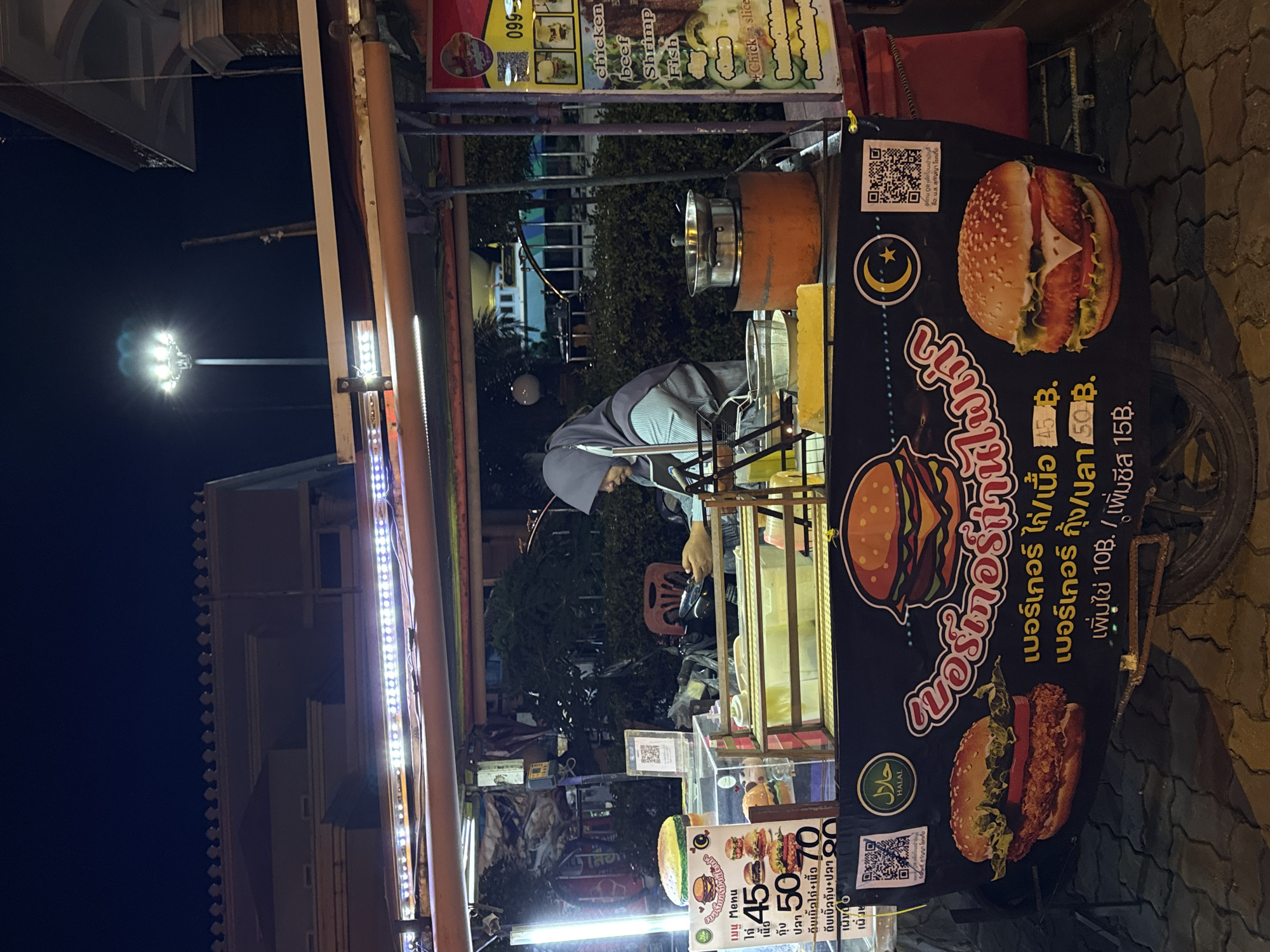 A street food cart is set up at night, likely in a city as indicated by the artificial lighting. The cart is decorated with illuminated strips of lights and features a large banner with images of burgers and Thai text. A person wearing a grey hijab and apron is operating the cart, seemingly preparing food or managing orders. Various food items and cooking equipment are visible on the cart, including pots and what appears to be a serving station. QR codes are displayed on the cart, suggesting a cashless payment option or a link to more information. The ground in front of the cart is paved with cobblestones. The background shows a building with architectural details and greenery, indicating an urban or suburban setting.