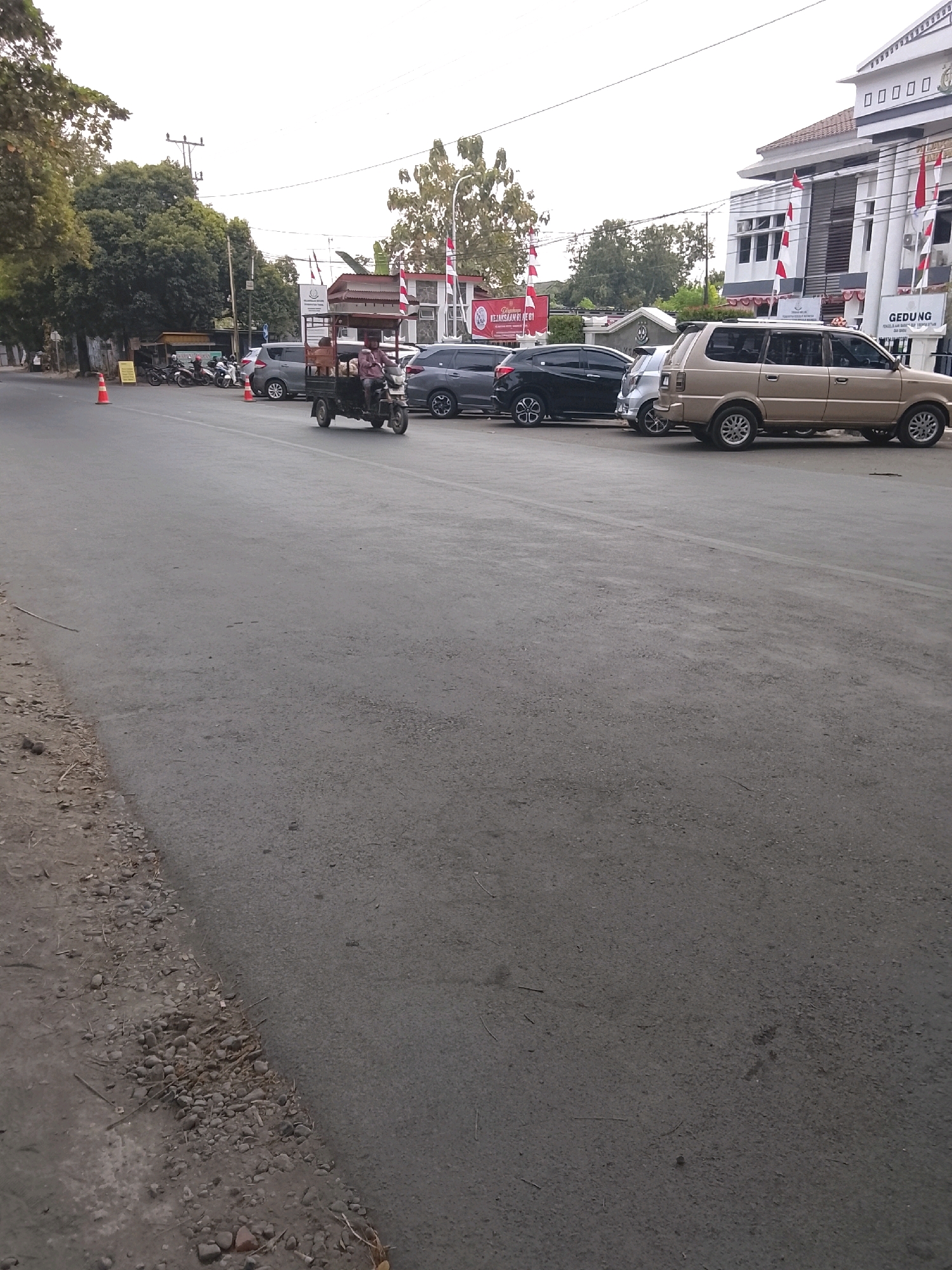 The image depicts a street scene in Slawi, Indonesia, likely in front of the Kejaksaan Negeri Slawi (Slawi District Attorney's Office). The foreground is dominated by a paved road with a gravelly shoulder. In the middle ground, a three-wheeled motorized vehicle carrying goods and two people is moving on the road. Several cars are parked along the side of the road, including a gold-colored SUV, a black sedan, and a silver hatchback. In the background, a multi-story building with a white facade, red and white flags, and signage indicating "GEDUNG" (Building) is visible. Trees line the street, and power lines are present. The sky is overcast.