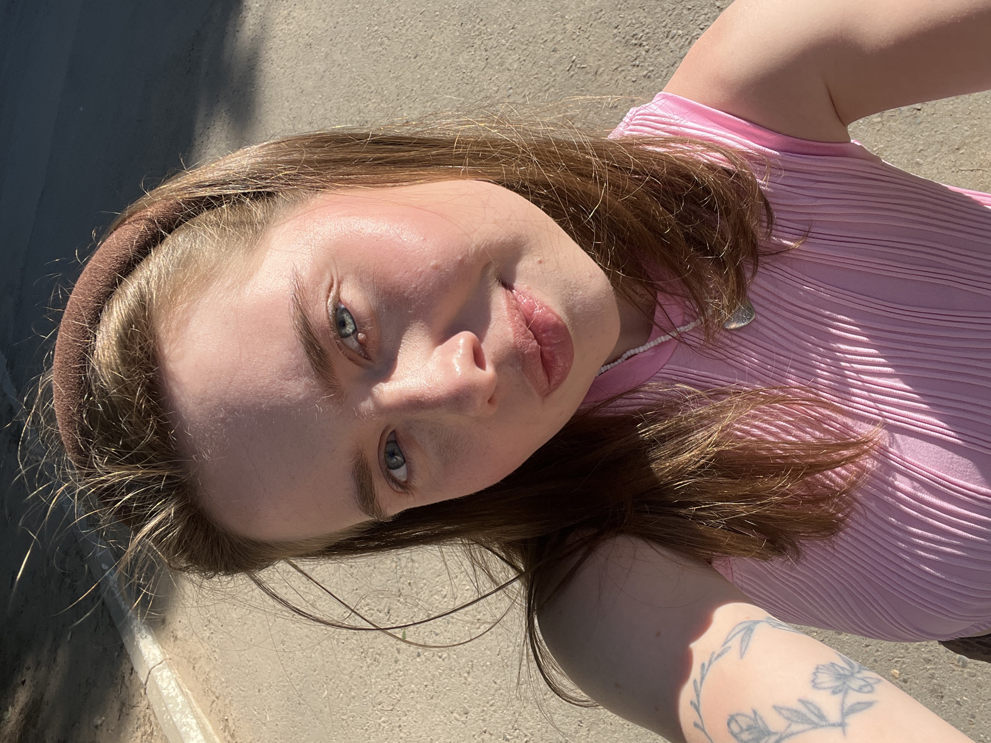 A young woman with light brown hair and blue eyes is pictured in a selfie, taken outdoors in Chaikovsky, Russia. She is wearing a pink ribbed top and a brown headband. Her left arm is visible, showing a tattoo of flowers and leaves. The background is a paved surface, possibly a sidewalk or road, with some shadows and sunlight. The lighting suggests it is daytime, possibly late afternoon due to the angle of the sun.