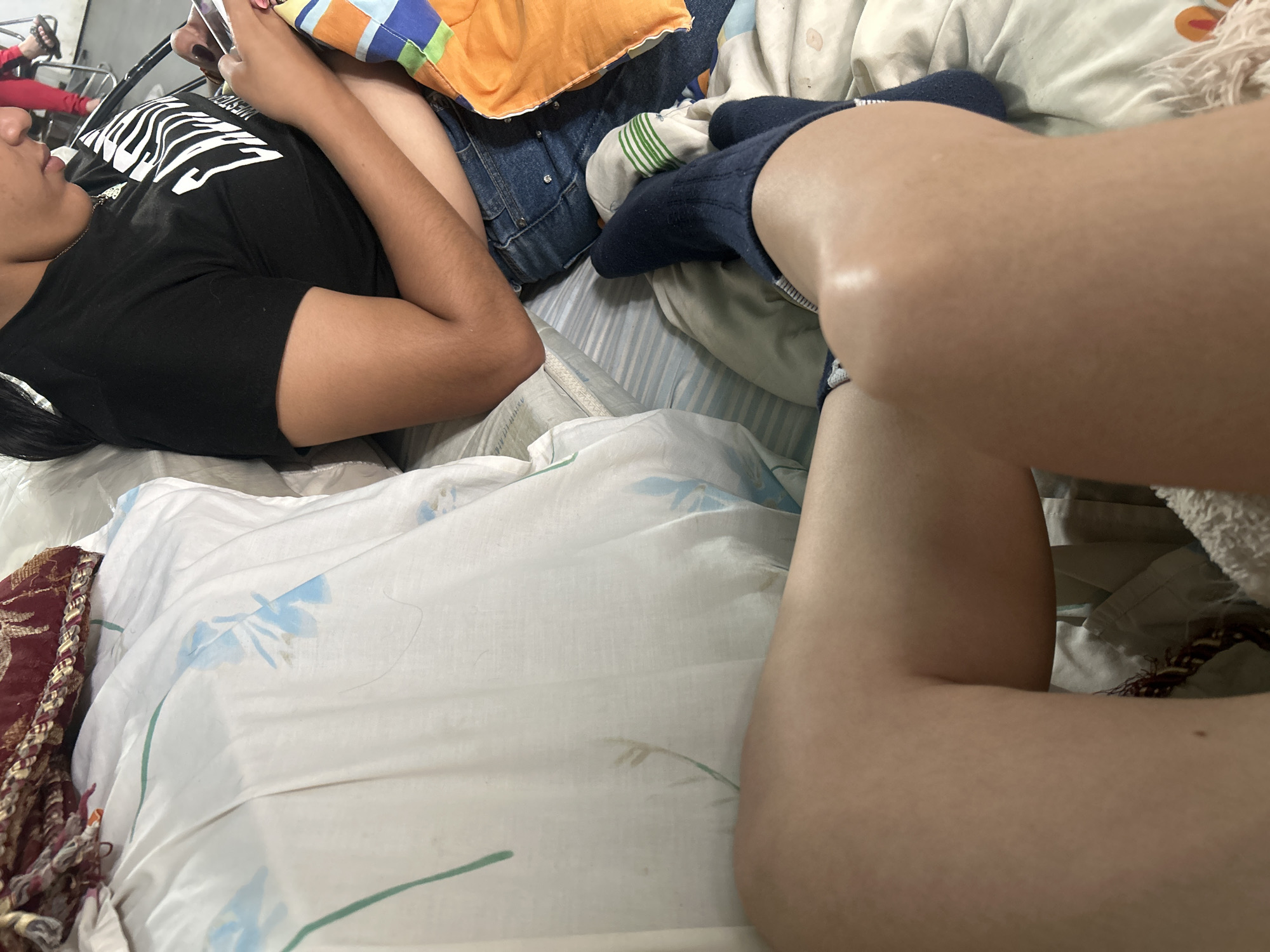 The image shows a close-up view of two individuals on a bed in what appears to be a bedroom. One individual, wearing a black t-shirt with white lettering and blue jeans, is seated and looking at a mobile phone. Their arm is resting on a colorful, striped blanket. The other individual's legs and knees are prominently featured in the foreground, wearing dark blue socks. The bed is covered with white and striped sheets. In the background, a portion of a maroon and gold decorative textile is visible. The overall scene suggests a casual, indoor setting, possibly a domestic space in Juan Griego, Venezuela.