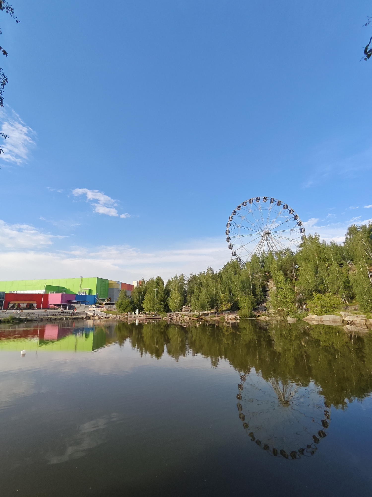 This is a photograph of the Ferris wheel at the "Uralmash Park" in Yekaterinburg, Russia, on a clear day. The Ferris wheel, a prominent amusement ride, stands tall amidst a line of green trees. In the foreground, a calm body of water reflects the trees, the Ferris wheel, and the colorful buildings of what appears to be a shopping center or entertainment complex on the left side of the frame. The sky is a vibrant blue with scattered white clouds. The scene depicts a recreational area with a mix of natural elements like trees and water, and man-made structures typical of a park or amusement venue.