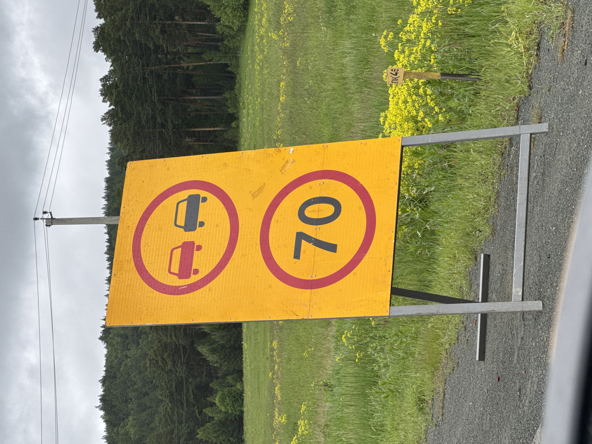 A bright yellow, rectangular traffic sign with rounded corners is the main subject of the image. The sign is supported by a metal A-frame structure. The top portion of the sign features a red circle with a red line through it, enclosing two car silhouettes. The left car is red, and the right car is dark gray. Below this, another red circle with a red line through it encloses the number "70" in black, indicating a speed limit. The sign is positioned on the side of a gravel road, with a grassy field dotted with yellow wildflowers to its right. In the background, a dense forest of pine trees stretches across rolling hills under a cloudy sky. Power lines and a utility pole are visible in the upper portion of the image. A small yellow marker with Cyrillic lettering and numbers "ПК 46" is partially visible in the field of wildflowers to the right of the sign. The scene appears to be in a rural or semi-rural area.