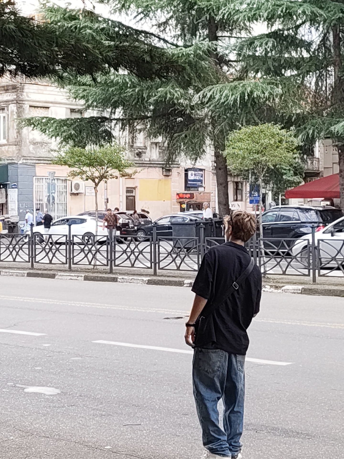 The image shows a person standing on a street in Batumi, Georgia, facing away from the camera. The person is wearing a black t-shirt, blue jeans, and sneakers, with a black bag slung across their chest. In the background, there are several cars parked along the street, including a white sedan and several black SUVs. Buildings with traditional architecture are visible behind the cars. Trees line the street, providing shade. A black metal fence separates the sidewalk from the street. The scene appears to be a public area in the city.
