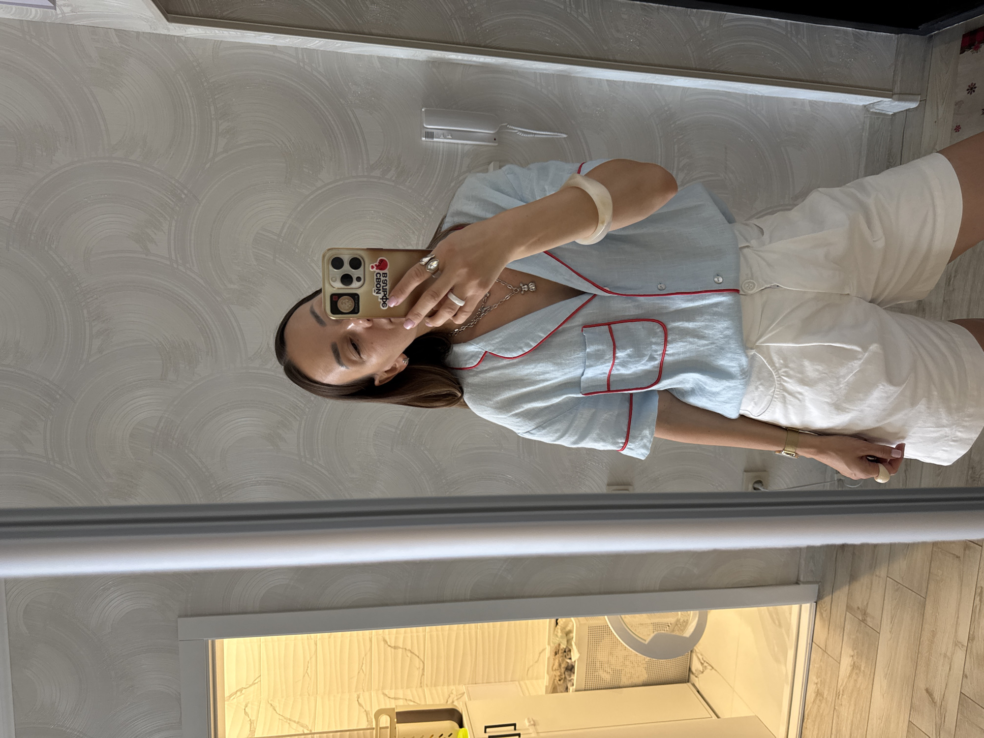 A woman takes a mirror selfie in a brightly lit room with textured wallpaper. She is wearing a light blue short-sleeved pajama-style top with red trim and white pleated shorts. She has on several rings and necklaces, and a wide, light-colored bracelet on her left wrist. In the reflection, a doorway leads to a bathroom with white tiled walls, a white washing machine, and a laundry basket. The floor is light-colored wood. The scene appears to be an indoor residential setting, possibly an apartment or hotel room. There is no visual information within the image to indicate the location is Sochi, Russia.