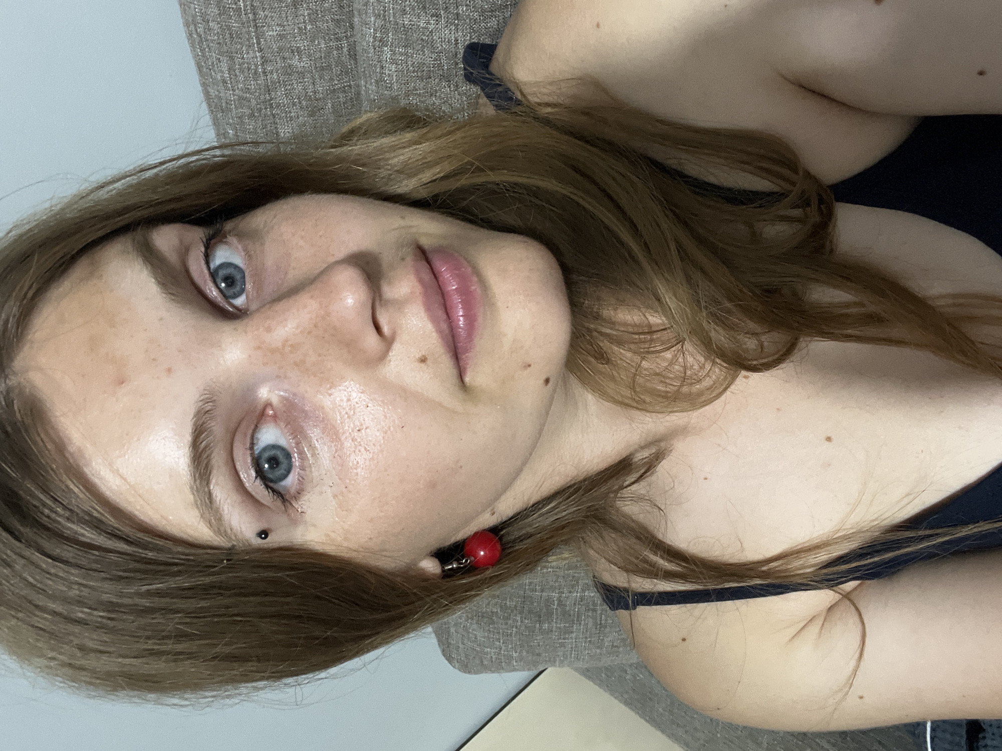 A young woman with light brown hair and blue eyes is pictured in a close-up, selfie-style shot. She has fair skin with freckles and a small piercing above her left eyebrow. She is wearing a dark blue tank top and a red cherry-shaped earring in her left ear. The background is a textured, light brown sofa and a pale blue wall, suggesting an indoor setting, likely a home. There is no discernible information to place this image in the city of Сеніцкі сельскі Савет, Belarus.