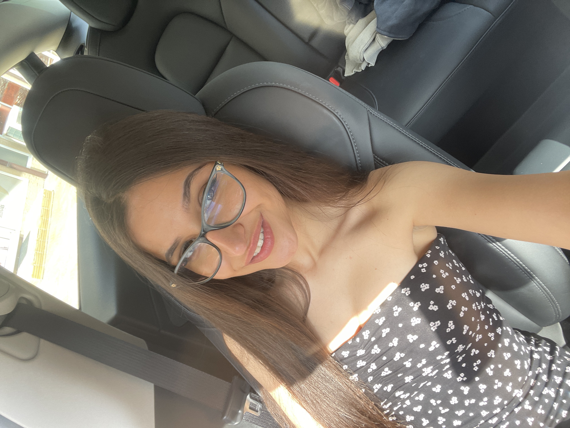 A young woman with long, dark hair and glasses is taking a selfie in the back seat of a car. She is wearing a black strapless dress with a white floral pattern. Sunlight is streaming in through the windows, casting shadows on her face and hair. The car's black leather seats are visible, as is a seatbelt. In the background, the outline of buildings and a street can be seen through the windows.