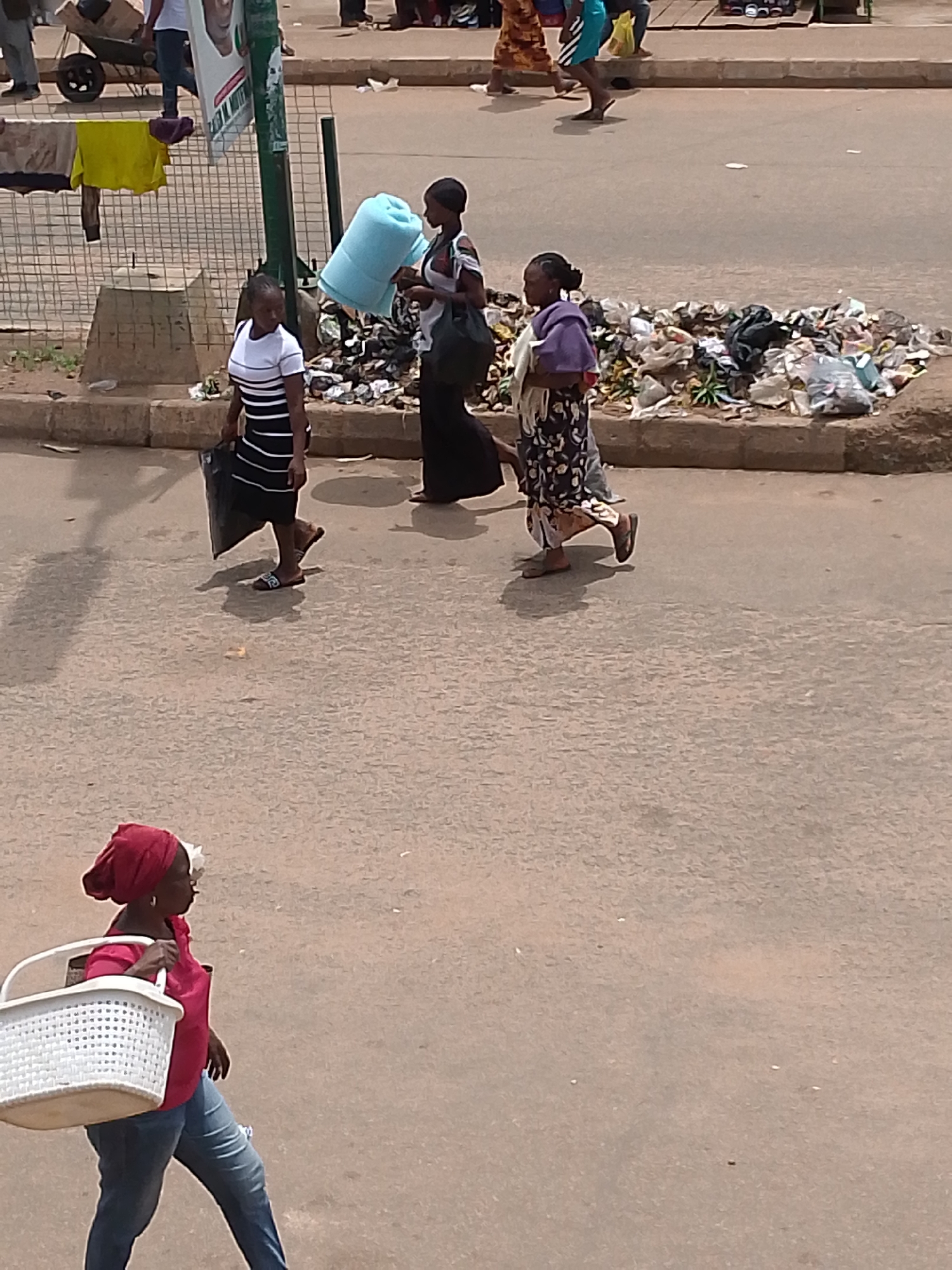 The image shows a street scene with several people. In the foreground, a woman wearing a red headscarf and a pink top, along with blue jeans, carries a white basket over her left shoulder. She is walking on a paved surface. In the midground, three women are walking. The woman on the left wears a black and white striped dress and carries a black bag. The woman in the middle, wearing a black dress, is holding a large, light blue object. The woman on the right wears a purple and white patterned dress and carries a bag over her shoulder. Behind the women in the midground is a pile of trash. In the background, there is a metal fence with yellow and purple clothing hanging on it, as well as a wheelbarrow. People are visible further in the background, some walking and others standing. The overall environment appears to be an outdoor urban setting.