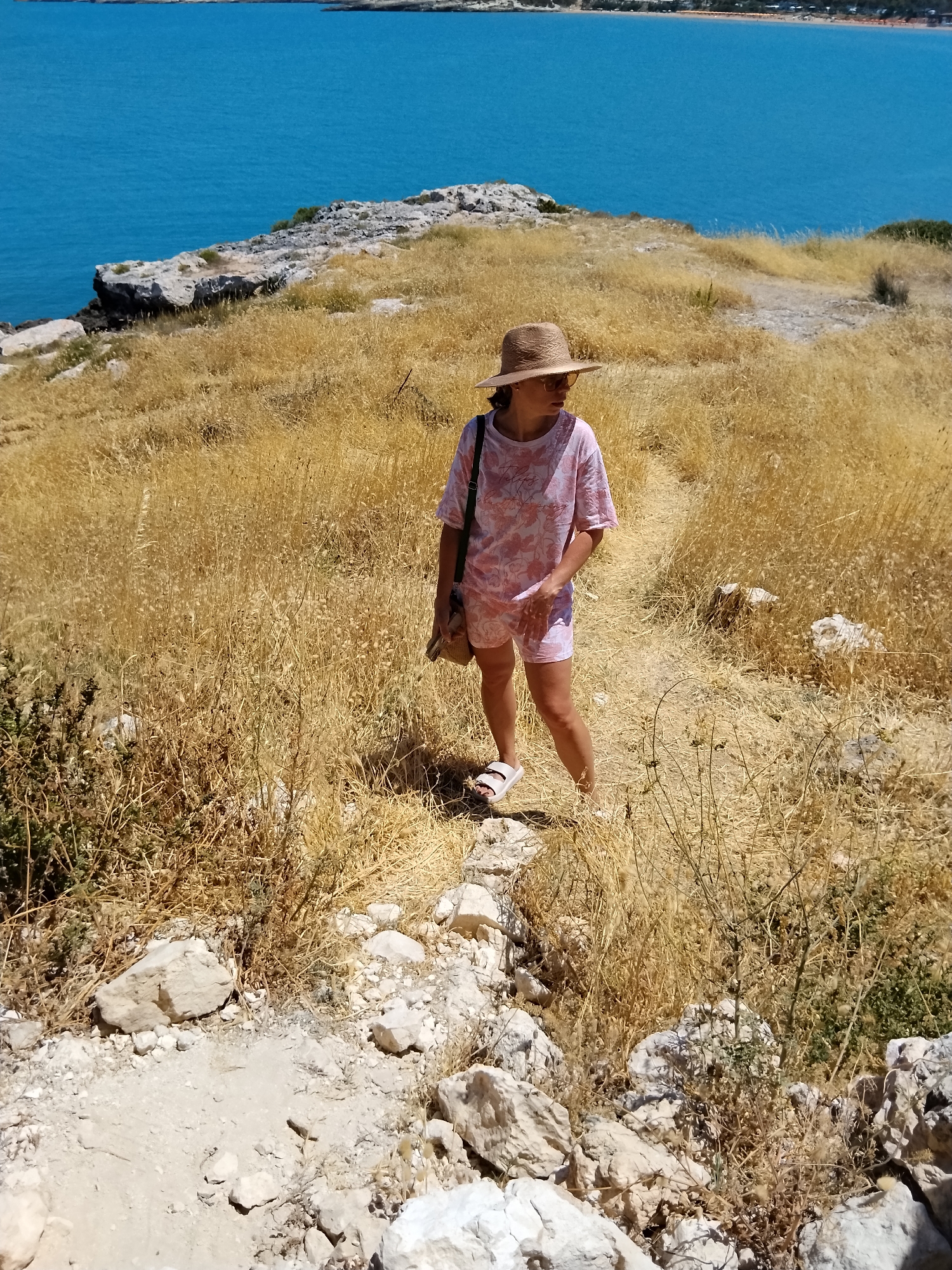 A person wearing a pink t-shirt, pink shorts, a straw hat, sunglasses, and white sandals stands on a dry, grassy hillside overlooking a bright blue body of water. The person is also carrying a small bag. The hillside is covered in golden-brown grasses and rocks. In the background, the vibrant blue water meets a distant shoreline with buildings. The scene appears to be a sunny day, likely in the late morning or early afternoon, given the bright sunlight and shadows. The overall impression is one of a scenic coastal landscape, possibly a hiking trail or viewpoint. The location is Peschici, Italy.