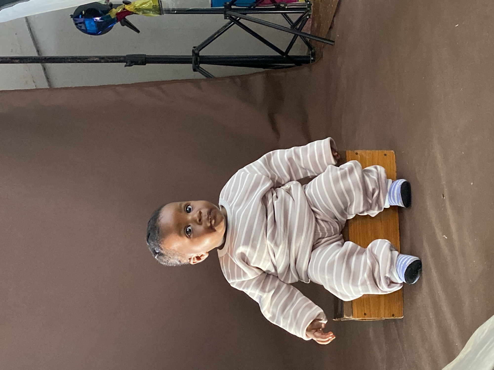 A baby wearing a striped pajama set and blue socks is sitting on a wooden surface. The background is a solid brown color. A black metal tripod or stand is visible in the upper left corner of the frame. The location cannot be ascertained from the image alone.