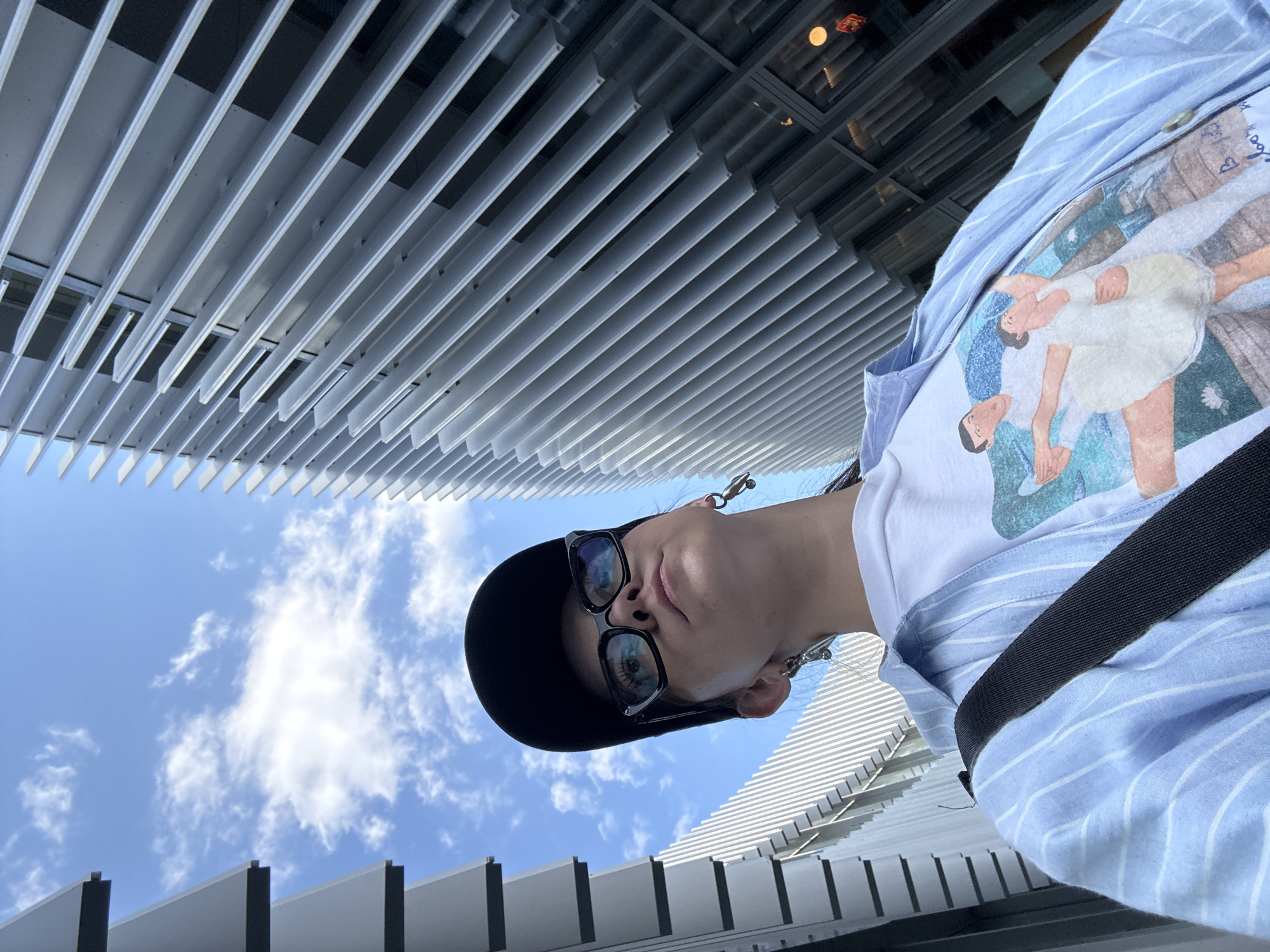 A person wearing a black hat, sunglasses, a white t-shirt with a graphic, and a blue striped shirt is looking down at the camera. The background includes a building with a facade of parallel, white horizontal slats, which contrasts with the blue sky and scattered clouds. The photo's perspective is from a low angle, looking upwards. The location is Kazan, Russia.