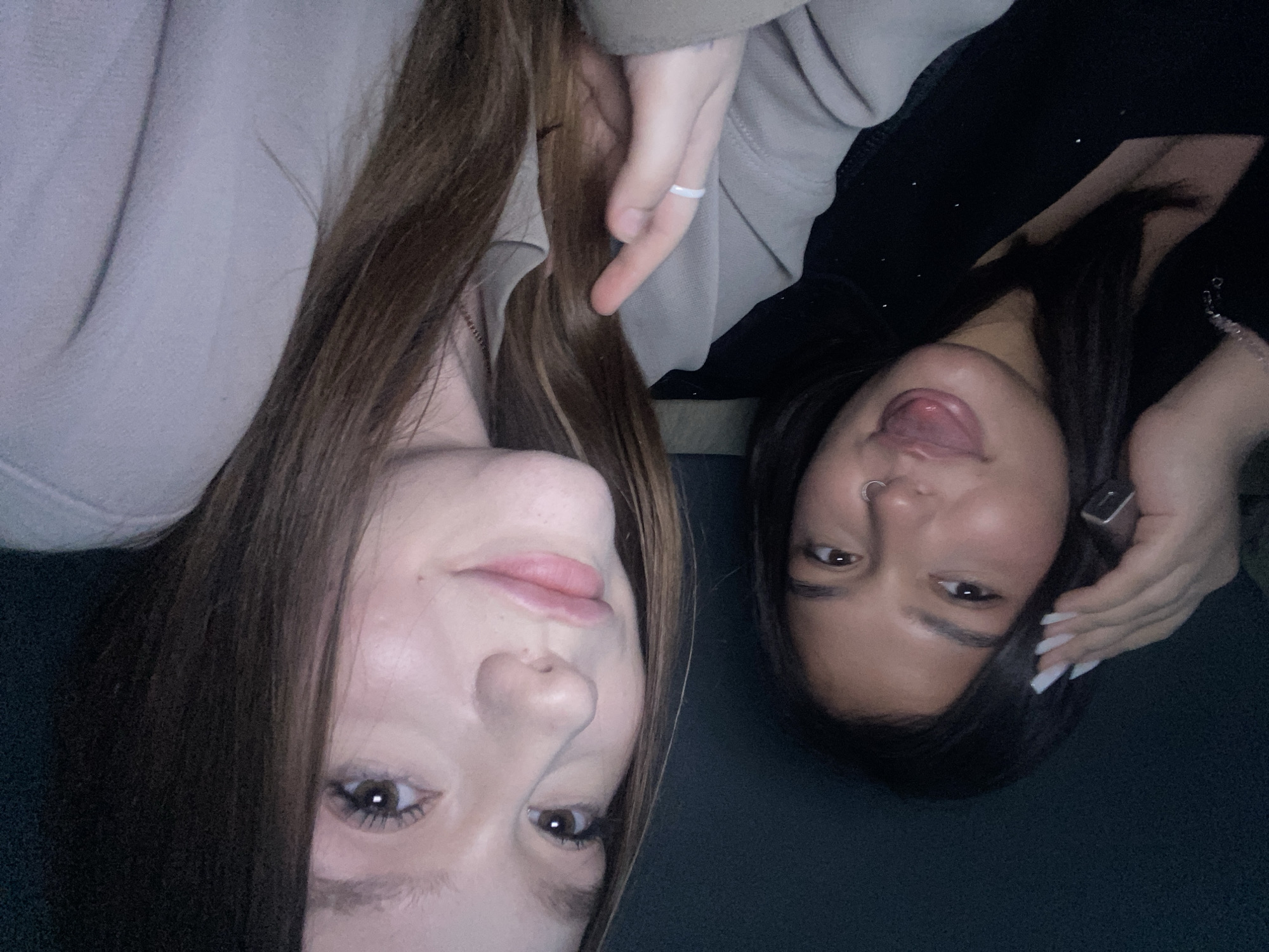 Two young women are posing for a selfie. The woman on the left has long brown hair and is wearing a light-colored sweatshirt. The woman on the right has dark hair and is wearing a dark top. She is sticking her tongue out and has a nose ring. The location cannot be determined from the image.