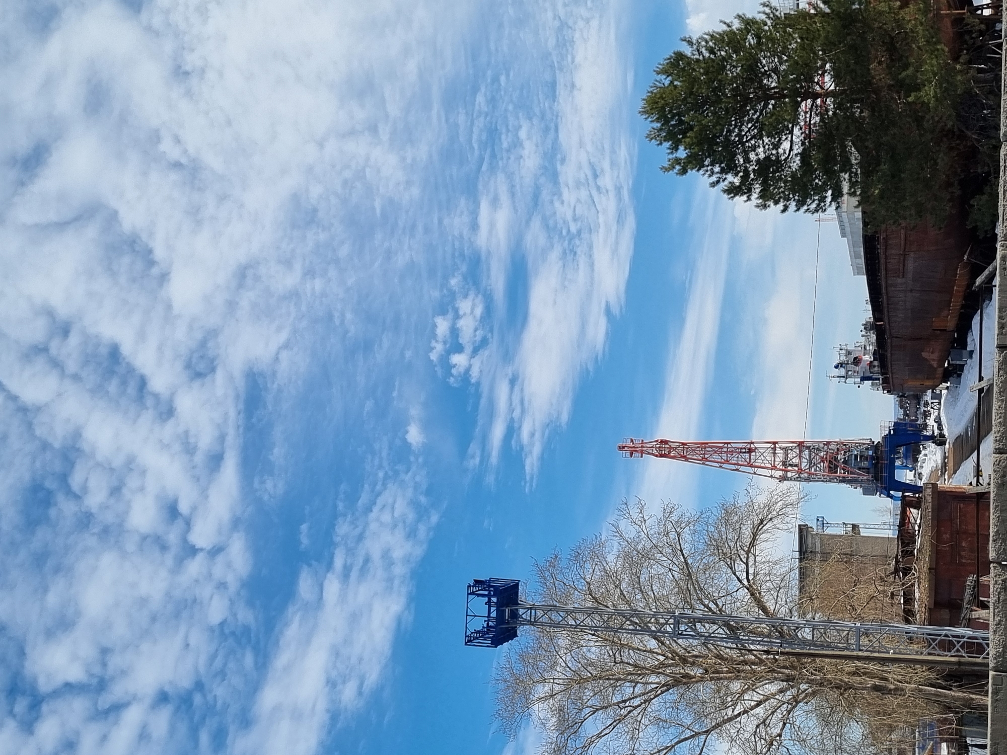 The image depicts an industrial river port or shipyard in Chaikovsky, Russia, under a partly cloudy blue sky. In the foreground on the left, a tall deciduous tree with bare branches stands next to a metal lattice tower topped with a blue square platform. Below the tree, a weathered, reddish-brown structure with a partially collapsed roof is visible. Further into the midground, a prominent red and blue gantry crane is positioned, suggesting cargo handling or shipbuilding operations. To the right of the crane, several large, dark, rusty barges or ship sections are situated on what appears to be a dry dock or repair slips, with patches of snow evident on the ground around them. A green coniferous tree stands on the far right, partially obscuring additional industrial elements or vessels in the background. The overall scene indicates a working industrial waterfront area during a cold season, characterized by industrial infrastructure, vessels undergoing maintenance, and wintry conditions. No individuals are discernible in the frame.