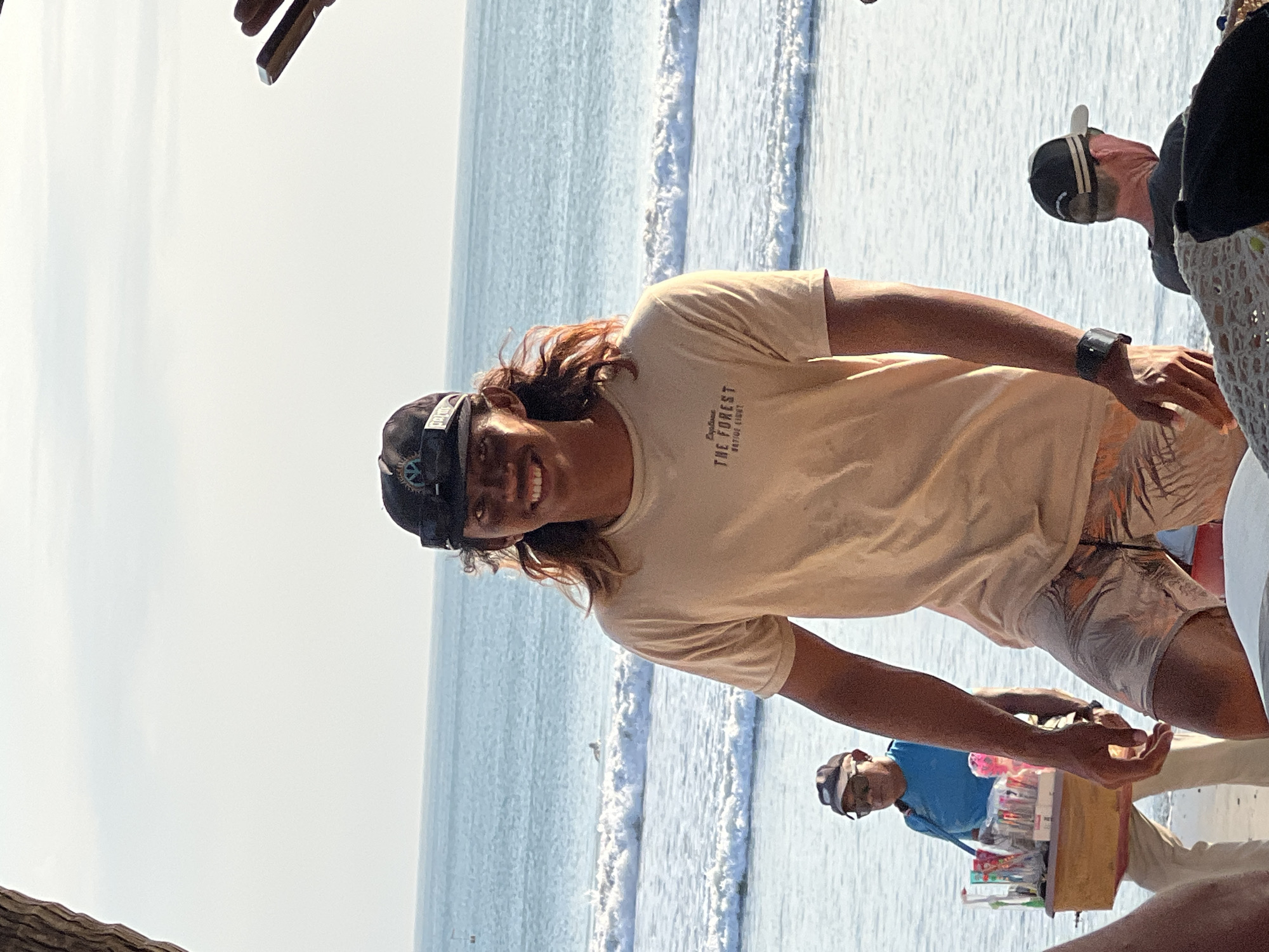 A young man with long, wavy brown hair and a tan complexion stands smiling at the camera on a beach. He wears a tan t-shirt with a logo that reads "THE FOREST" and a black baseball cap. He has on patterned shorts and a black watch. In the background, the ocean with gentle waves is visible, and a surfer can be seen in the distance. To the left, a person wearing a blue shirt and sunglasses walks by with a wooden cart filled with various items. Further to the right, another person, seen from the back, sits on the beach wearing a baseball cap. The sky is clear with a light haze. The scene appears to be a warm, sunny day in Kuta, Indonesia.