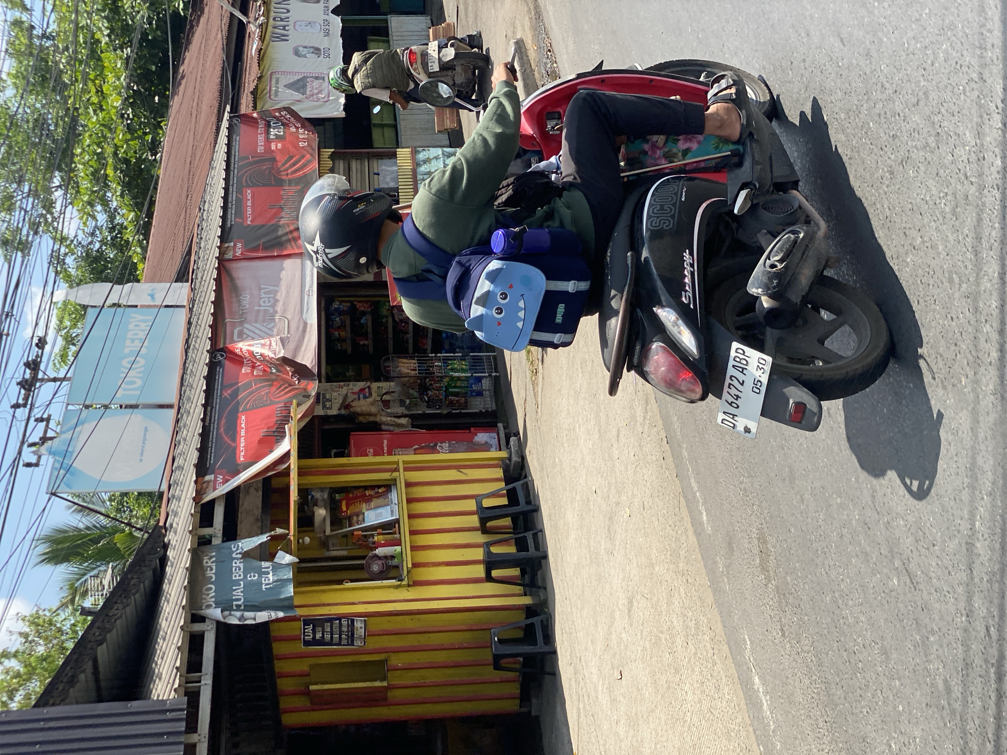 The image depicts a street scene in Banjarmasin, Indonesia. In the foreground, a person wearing a helmet and a green long-sleeved shirt is riding a black scooter. The rider has a blue backpack with a cartoon monster design on it, and a blue water bottle attached. The scooter has a license plate that reads "DA 6472 ABP 05-30". The scooter is passing by a row of small shops. One shop, painted yellow and red, has signs advertising "JUAL BERAS & TELUR" (Selling Rice & Eggs) and "JUAL PULSA" (Selling Phone Credit). Another shop displays a menu and what appears to be a Coca-Cola refrigerator. Further down the street, another person on a scooter is visible, and a sign for "WARUNG SOTO NASI SOP AYAM PA" is present. The environment is sunny with trees and power lines overhead. The road surface is asphalt.