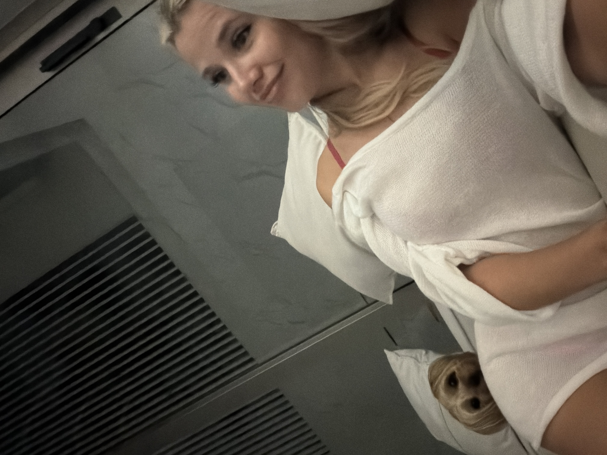The media file shows a close-up, high-angle shot of a young woman with blonde hair, wearing a white terrycloth robe over a red bikini top. She is looking down and smiling, with her hair falling around her face. A small dog with blonde fur is peeking out from under the robe, nestled on a white pillow. The setting appears to be indoors, possibly a hotel room or spa, with a ribbed wall or furniture in the background. The lighting is soft and diffused, suggesting it might be morning or evening. The overall mood is relaxed and intimate. There is no visible text in the image.